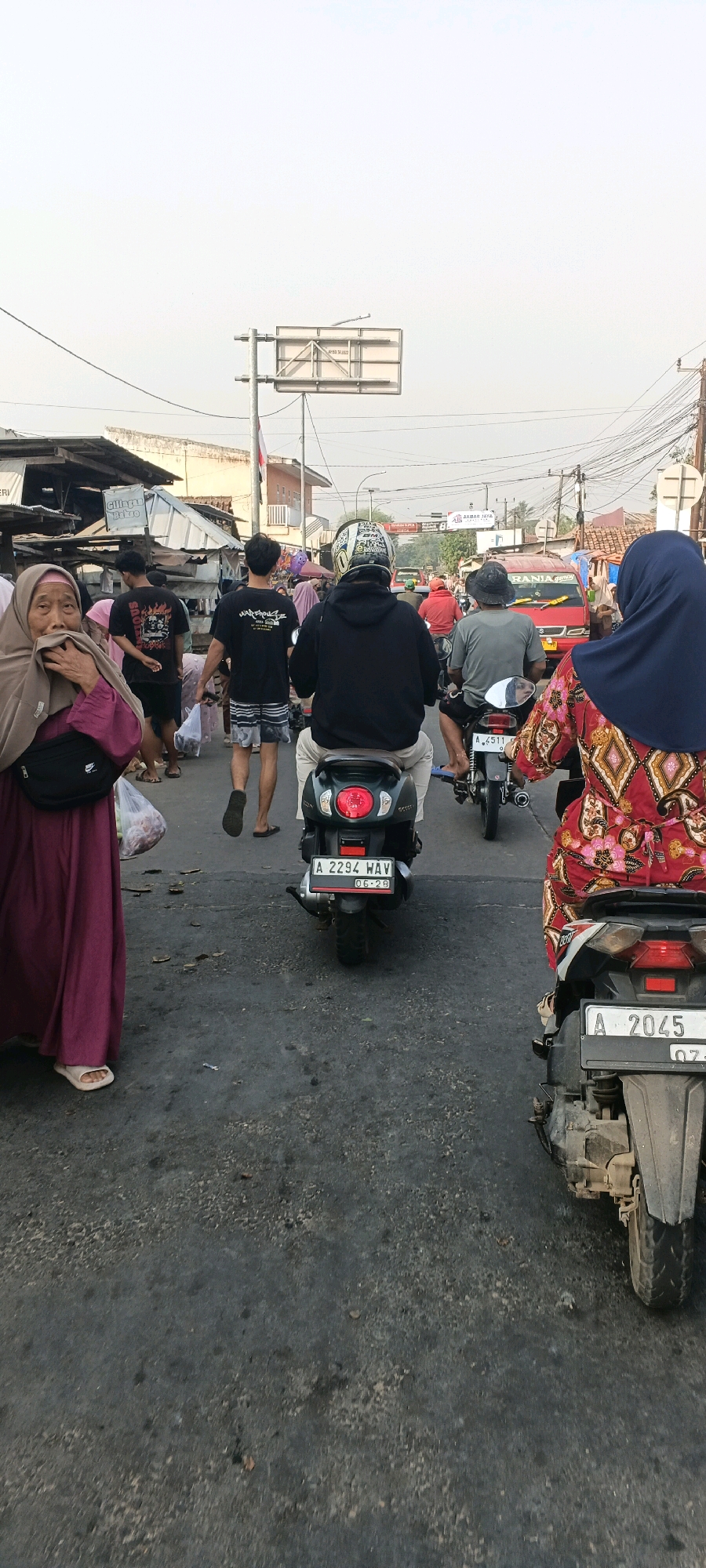 The image depicts a busy street scene in Singaraja, Indonesia, likely near a market or commercial area. Several individuals are on foot and on motorcycles. On the left, a woman wearing a purple hijab and a maroon dress is walking and holding a plastic bag. Behind her, several men are walking, one in a black t-shirt with graphics and another in a striped t-shirt. In the center, a person wearing a black hoodie and helmet rides a dark green scooter with license plate "A 2294 WAV". To the right, a woman in a colorful, patterned dress and dark blue hijab is on a scooter. Further back, a red van is visible in traffic. The environment appears to be a road with buildings and stalls lining the sides. Overhead, there are electrical wires and a traffic sign. The overall impression is of a lively, somewhat congested urban setting.