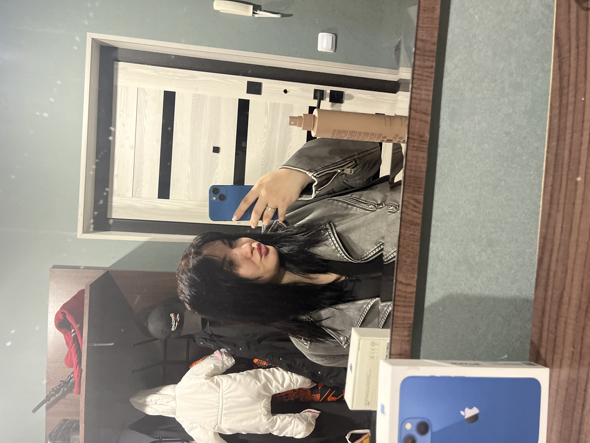 The image shows a selfie taken in a mirror in what appears to be a domestic setting. A young woman with black hair, wearing a grey jacket, holds a blue phone. A beige spray bottle is visible in the upper part of the mirror. Reflected in the mirror are objects on a dark-colored surface, including a white baby snowsuit, a black baseball cap, and an iPhone box. A door with a light frame and dark inset panels is visible in the background. The walls appear to be painted a light green. The location, Chaikovsky, Russia, is not discernible from the image.