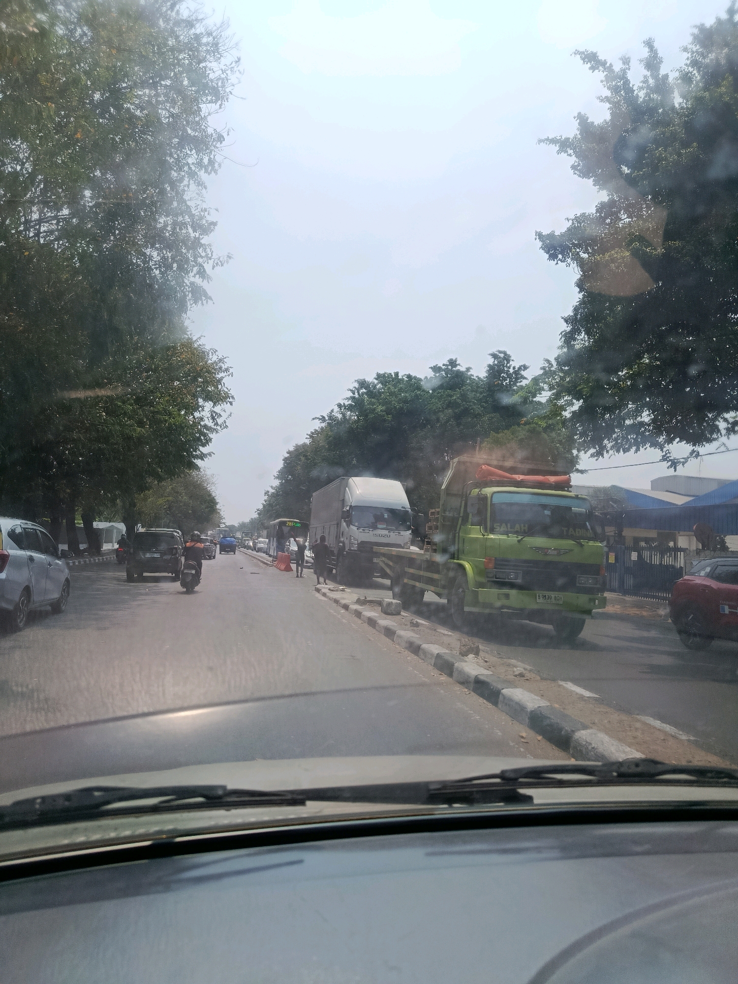 A slightly hazy, overhead shot captures a street scene in East Jakarta, Indonesia, likely on a bright, possibly humid day. The foreground is dominated by a car's dashboard and windshield wipers, suggesting the photo was taken from inside a moving vehicle. The road, paved and in moderate condition, shows signs of light to moderate traffic. On the right side of the road, a green dump truck with the word "SALAH" visible on its windshield is parked alongside a row of buildings with blue and white awnings. Further down the road, a white truck with "ISUZU" on its side and a red car are visible. On the left, a silver car is parked, and beyond it, trees line the street, their foliage dense and green. A few individuals are seen on the side of the road, one near the green truck and another further down near a bus. The sky is bright and appears overcast or hazy.