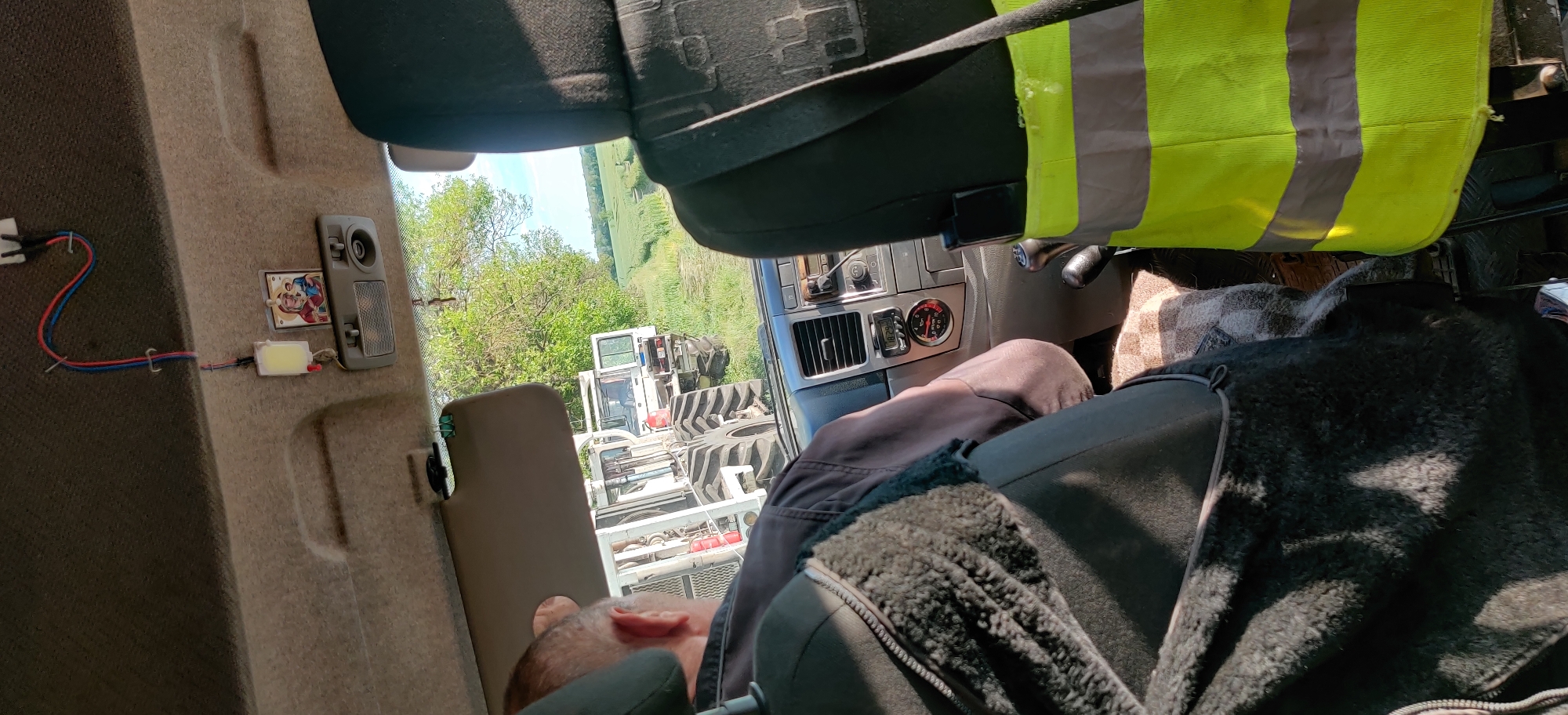 The image shows the interior of a vehicle, possibly a tractor, parked in a field near Randava, Ukraine. A man is seated, partially obscured by the seat, wearing a black jacket with a sheepskin-like lining. A high-visibility vest is draped over the passenger seat. Visible through the window is another piece of machinery, appearing to be a tractor or agricultural equipment. The vehicle's dashboard includes a digital display showing the time as 12:18. The vehicle's interior includes a light fixture and an attached sticker. A red and blue wire is attached to the fixture. The surrounding environment features green fields and trees.