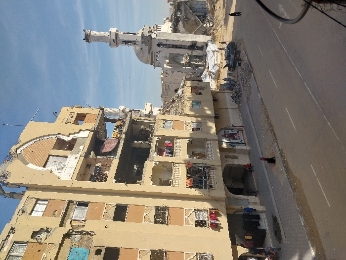 The image captures a street scene in Khan Yunis, Palestinian Territory, characterized by significant structural destruction under a clear blue sky. On the left, a multi-story, beige-colored building, likely residential or commercial, exhibits extensive damage, with upper floors largely collapsed or exposed, revealing interiors and rebar. A large arched facade element on its upper level is partially destroyed. Lower floors show broken windows and balconies, some with clothes hanging. Debris is accumulated at the base of this building and on the adjacent sidewalk. To the right, a white mosque with a dome and a prominent minaret also displays damage, surrounded by significant rubble and partially collapsed structures, some covered by white tarps. A paved two-lane road with white markings spans the foreground. Several individuals are present: a few people stand on the sidewalk near the damaged building, a small group including children are visible under the arches of the building's ground floor, and a lone figure walks away from the camera on the street. Near the mosque, a dark-colored vehicle is parked on the road, with its hood possibly open or damaged, surrounded by a small group of people. Wires are partially visible in the lower right corner of the frame.