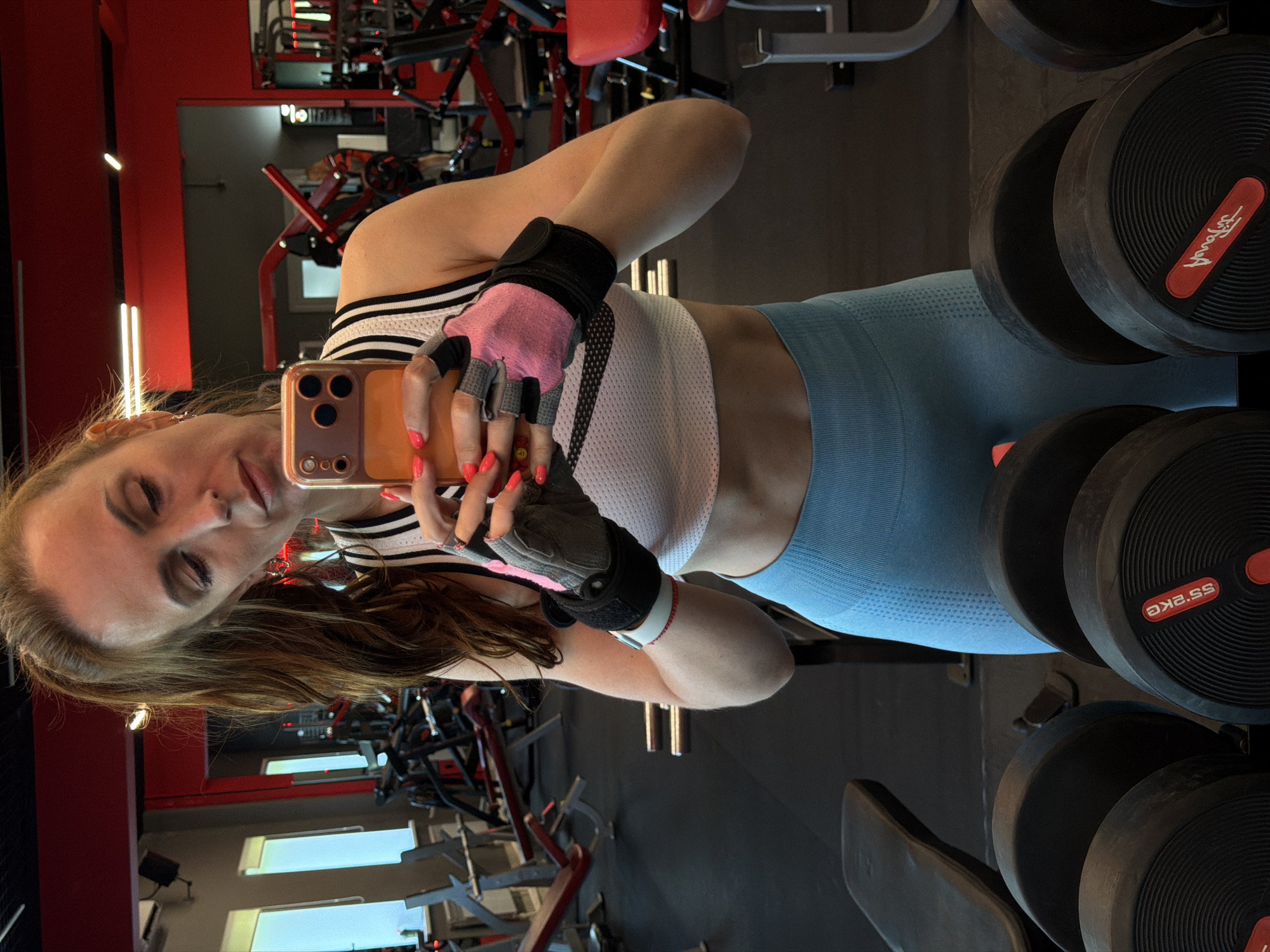 The image shows a woman taking a selfie in a gym. She is wearing a white sports bra, blue leggings, and lifting gloves. Her hair is tied back in a ponytail. She is holding an orange smartphone, and her fingernails are painted neon pink. In the background, there are various gym equipment, including weight machines and dumbbells. The gym appears to be well-lit with red accents on the walls and ceiling. The presence of a variety of exercise machines suggests it is a fully equipped fitness facility. The woman's posture and attire indicate she is actively engaged in or has recently completed a workout. The overall environment is that of a modern fitness center. The location is not identifiable as Birobidzhan, Russia, as no specific landmarks or textual clues indicate this location.