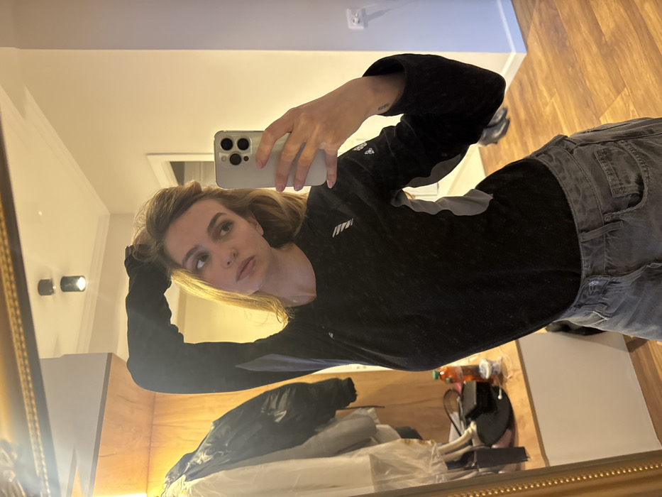 A female subject is depicted taking a mirror selfie indoors. She has light brown hair, which she is touching with her raised left hand. She wears a black long-sleeved athletic-style top with grey side panels and grey denim shorts. A small dark tattoo is visible on her right inner wrist. She holds a light-colored smartphone, equipped with a triple camera module, in her right hand, positioned to capture her reflection in the mirror. Her gaze is directed towards the smartphone.
The subject is reflected in a large, rectangular mirror framed in gold-colored ornate trim. The immediate environment, viewed in reflection, includes a light-colored wall on the right, featuring a white electrical outlet and a white doorframe in the distance. The floor is covered with wooden planks, and two dark shoes are visible on the floor beyond the subject. On the left side of the reflection, a wooden paneling or furniture piece is visible alongside a clothing rack holding several garments, including a white and a black jacket. Below the clothing rack, a wooden shelf or console displays a dark bag, a reddish-orange bottle, and other miscellaneous items. The ceiling, also reflected, is light-colored and features two dark, cylindrical spotlights. The location is an interior room within the city of Unknown, Unknown.