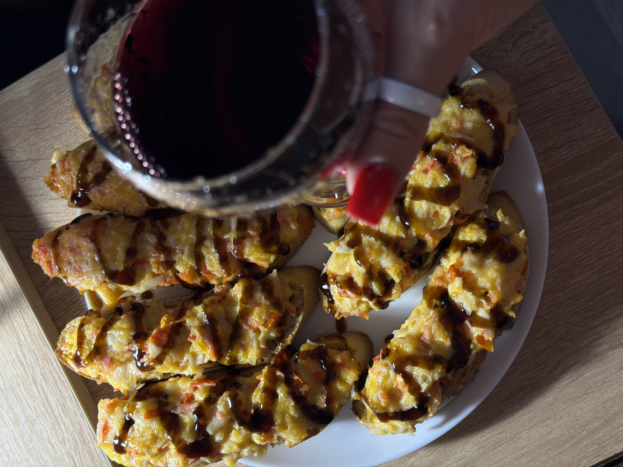 A person is holding a glass of red wine over a plate of open-faced sandwiches. The sandwiches are made with toasted bread topped with a mixture of cheese, some type of shredded meat or fish, and red flecks that could be bell peppers or crab. They are drizzled with a dark sauce, possibly balsamic glaze or teriyaki. The plate is white and rests on a wooden surface. The lighting suggests it might be daytime, with shadows cast by the wine glass and the plate, indicating natural light from the side. The red wine in the glass is a deep, rich color. The person's hand, with bright red nail polish, is visible as they hold the glass. The scene appears to be set indoors, likely in a dining area or kitchen, given the wooden table. The overall impression is one of a casual meal or appetizer enjoyed with a drink.