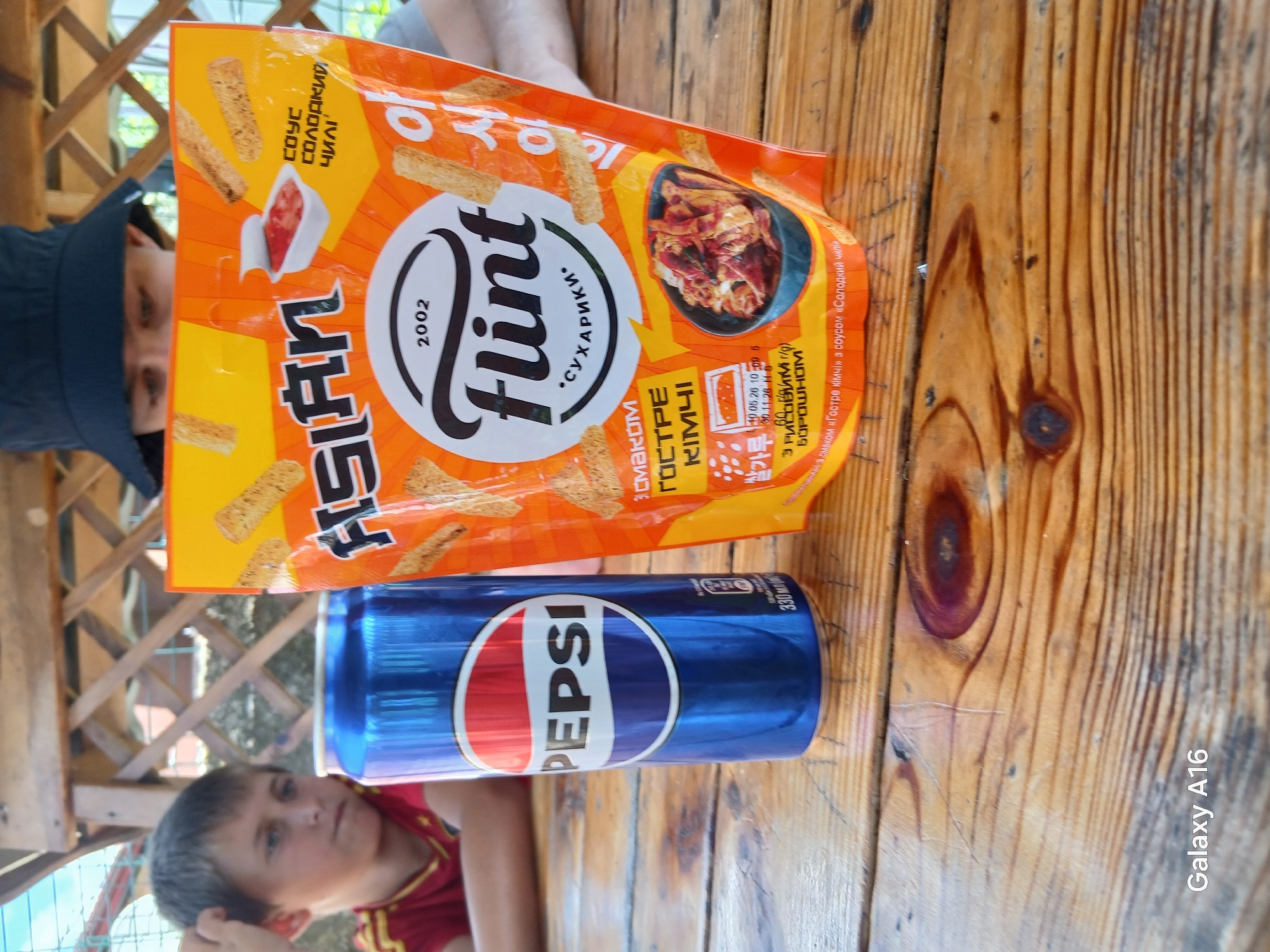The photograph captures a close-up view of a blue Pepsi can and an orange bag of Flint brand "suhariki" (croutons or crackers) placed on a wooden table, likely outdoors under a gazebo. A young boy, with dark hair and wearing a red shirt, is partially visible in the background, looking towards the right. To the right of the boy, a person wearing a dark hat is also visible in the background, partially obscured by the snack bag. The snack bag features an image of kimchi and text in what appears to be Ukrainian and possibly Korean, advertising a "spicy kimchi" flavor with "chili sauce." The Pepsi can shows its logo prominently. The wooden table has a visible grain and appears worn. The scene suggests a casual outdoor gathering or snack break. The "Galaxy A16" watermark in the bottom left corner indicates the device used to take the photo. The location of Ustyvytsia, Ukraine, is provided without any visual confirmation in the image itself, but if this were an annotation, it would be considered a factual detail.