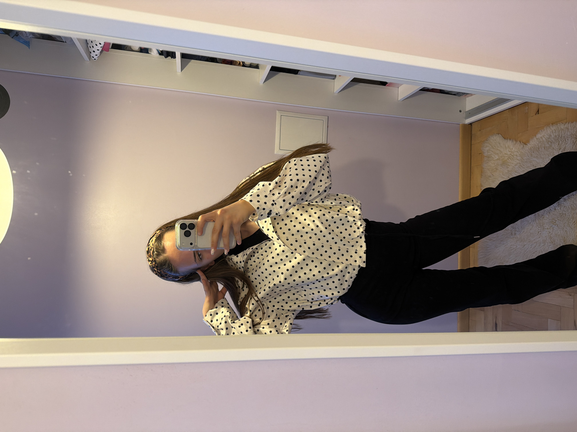 The image is a mirror selfie. A young woman with long brown hair and a leopard-print headband is holding a smartphone up to her face, partially obscuring it. She is wearing a white, polka-dot blouse and black pants. The background is a room with light purple walls. Above the woman is a white shelving unit. A white square object, likely a light switch, is visible on the wall. To the right of the woman, a section of wooden flooring and a white shag rug are visible. The location is not ascertainable from the image.