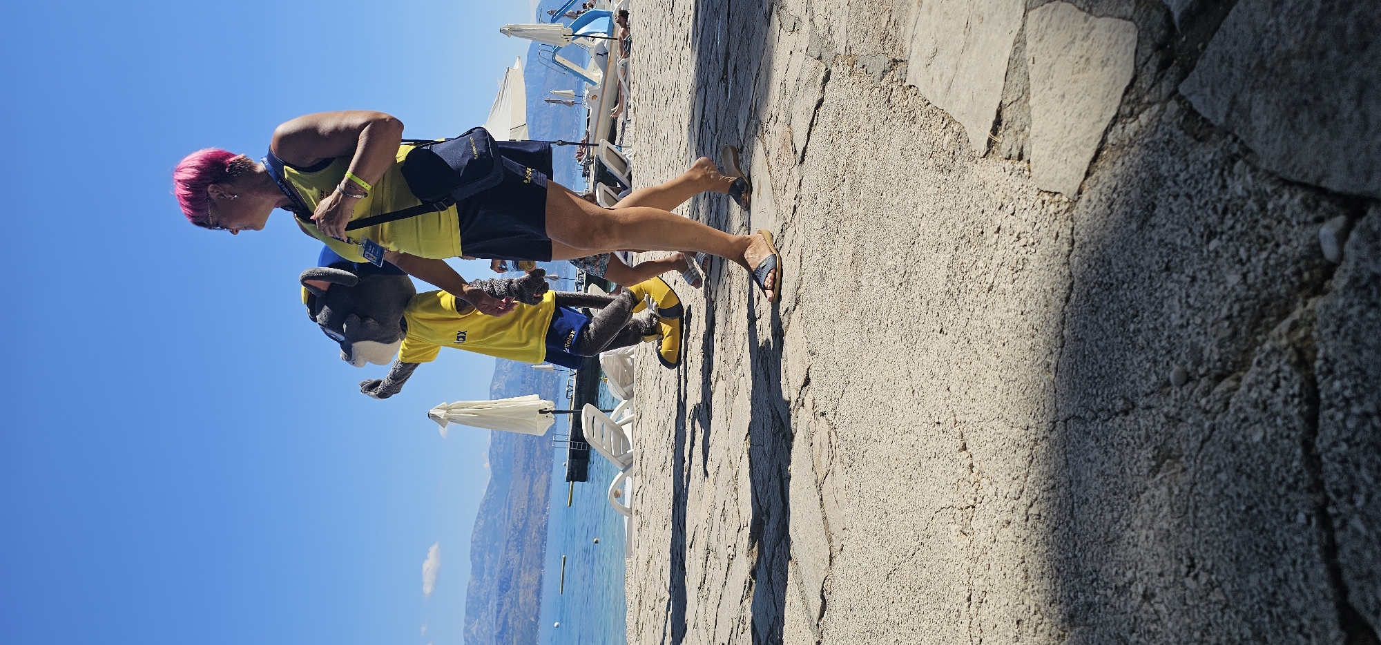 In Grad Supetar, Croatia, on a sunny day, a person with bright pink hair walks alongside a costumed character resembling a grey wolf wearing a yellow shirt and blue shorts. The person is wearing a yellow tank top and blue shorts and carries a dark blue bag over their shoulder. They are walking on a paved area next to the sea, with mountains visible in the distance. Several white beach umbrellas and lounge chairs are set up on the beach, and the water appears calm. The ground is made of rough, light-colored flagstones. The wolf mascot is waving one hand, and the person is holding onto the mascot's arm.