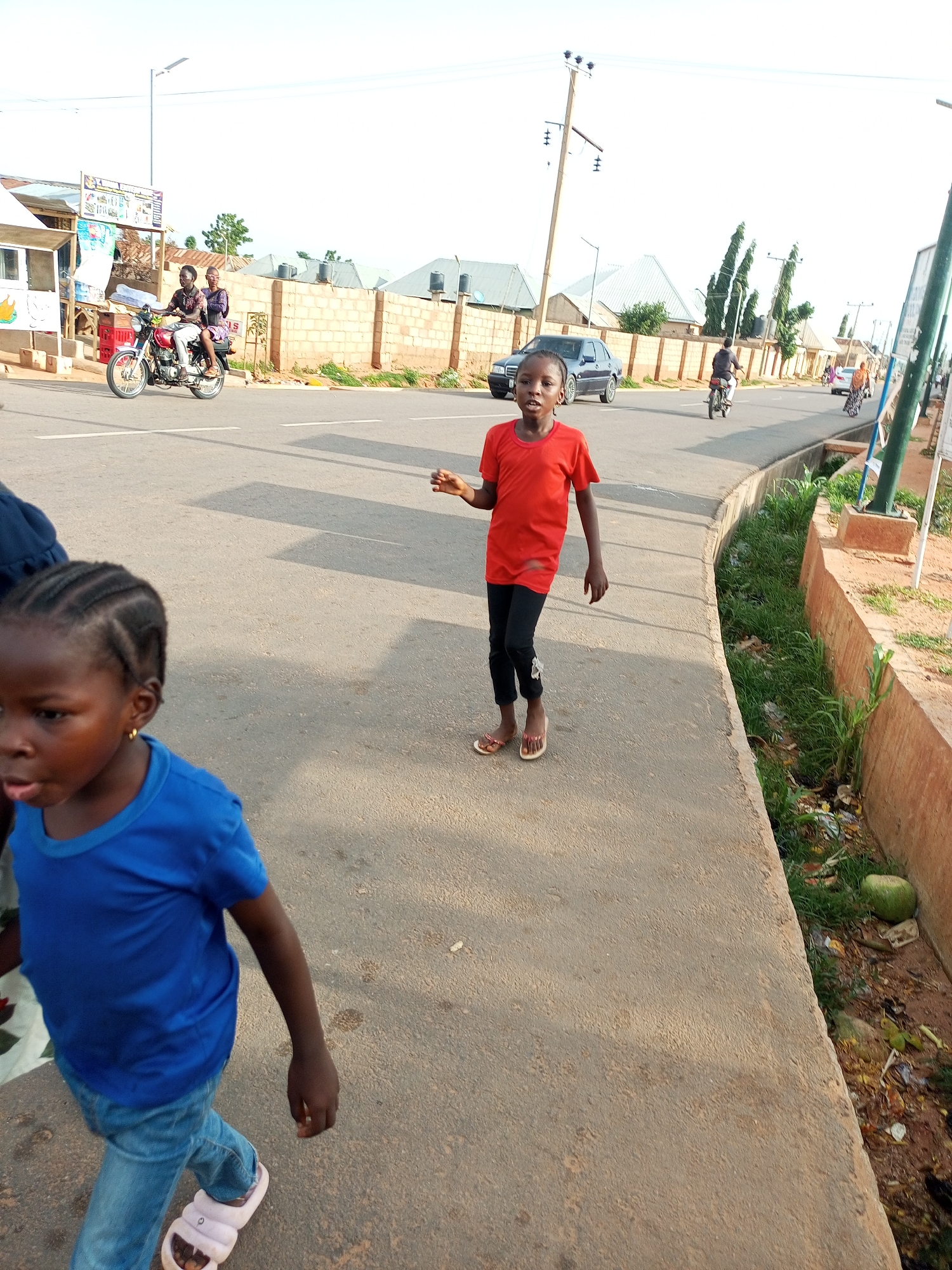 A street scene in Tumfure, Nigeria, captures children and vehicles on a sunny day. In the foreground, a young girl with dark braided hair, wearing a blue t-shirt and denim jeans, walks towards the left side of the frame. To her right, a slightly older child in a red t-shirt and black pants stands on the pavement, looking towards the viewer with an open mouth. Further down the street, a motorcycle with two occupants travels away from the camera. On the left side of the road, a small building with a white facade and a sign is visible, with a parked motorcycle in front of it. The street is paved, with a concrete drainage ditch running along the right side of the pavement, filled with overgrown grass and debris. The background shows a brick wall, houses with corrugated roofs, and utility poles. The sky is bright and clear.