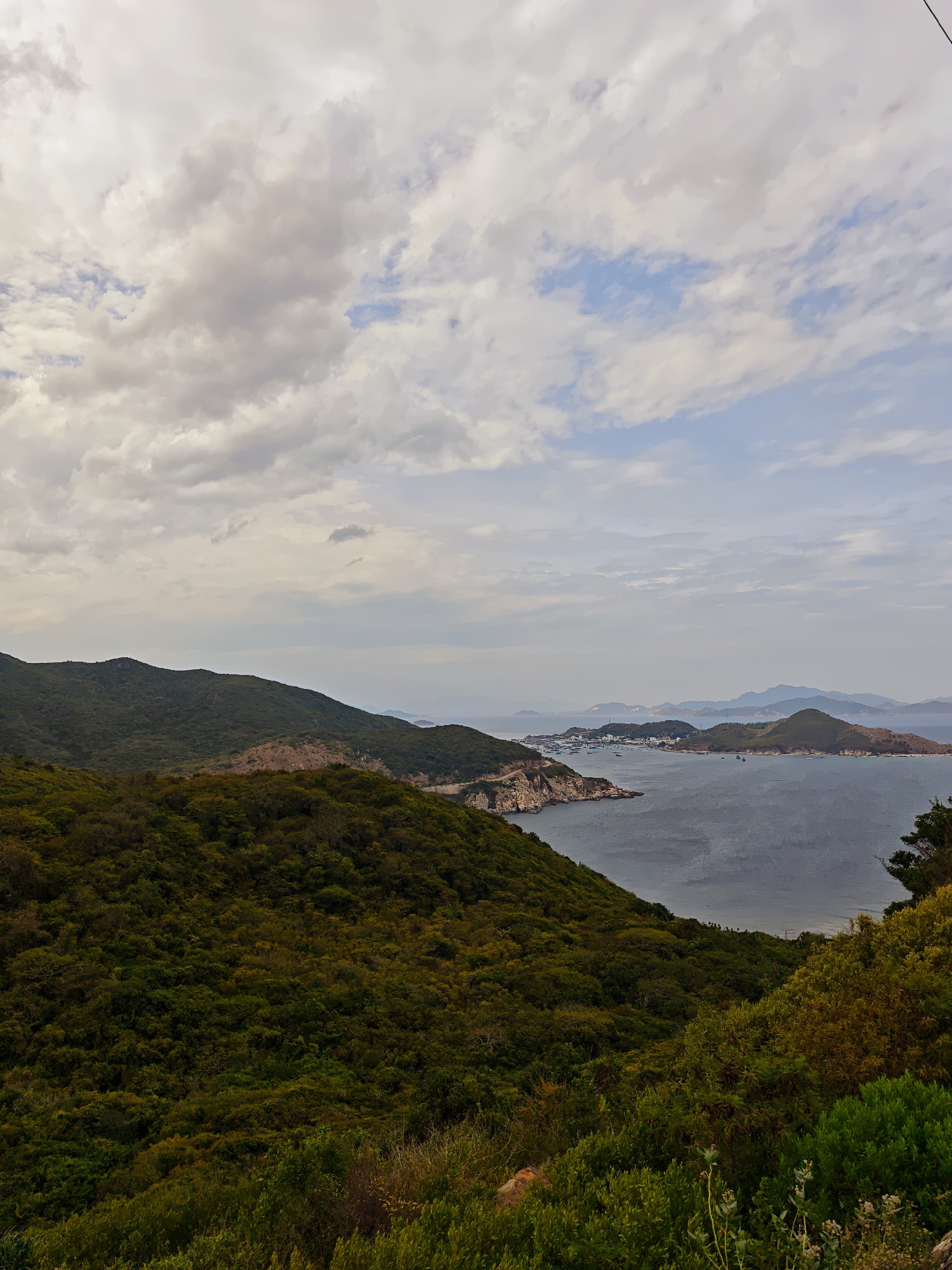 This is a high-angle, wide shot photograph taken from a hillside overlooking a bay with a town and several islands. The sky is overcast with scattered white and gray clouds, with patches of blue sky visible. The sea is a deep gray-blue.
In the foreground, there is dense, dark green vegetation covering the sloping hillside. The vegetation consists of various types of trees and bushes.
In the mid-ground, the bay curves around a peninsula with a town situated along its coast. The town appears to be a collection of buildings with some infrastructure, including what looks like a road winding along the coastline of the peninsula. Several boats are docked in the bay, and others are scattered across the water. Beyond the town, several islands of varying sizes are visible in the distance, some with green, hilly terrain.
The background consists of more distant islands and a hazy horizon where the sea meets the sky. The overall impression is of a scenic coastal landscape with human settlement integrated into the natural environment. The specific location in Vĩnh Hải, Vietnam, is not explicitly identifiable from the visual elements alone, but the landscape suggests a tropical or subtropical coastal region. There are no discernible people or specific actions taking place in the scene.