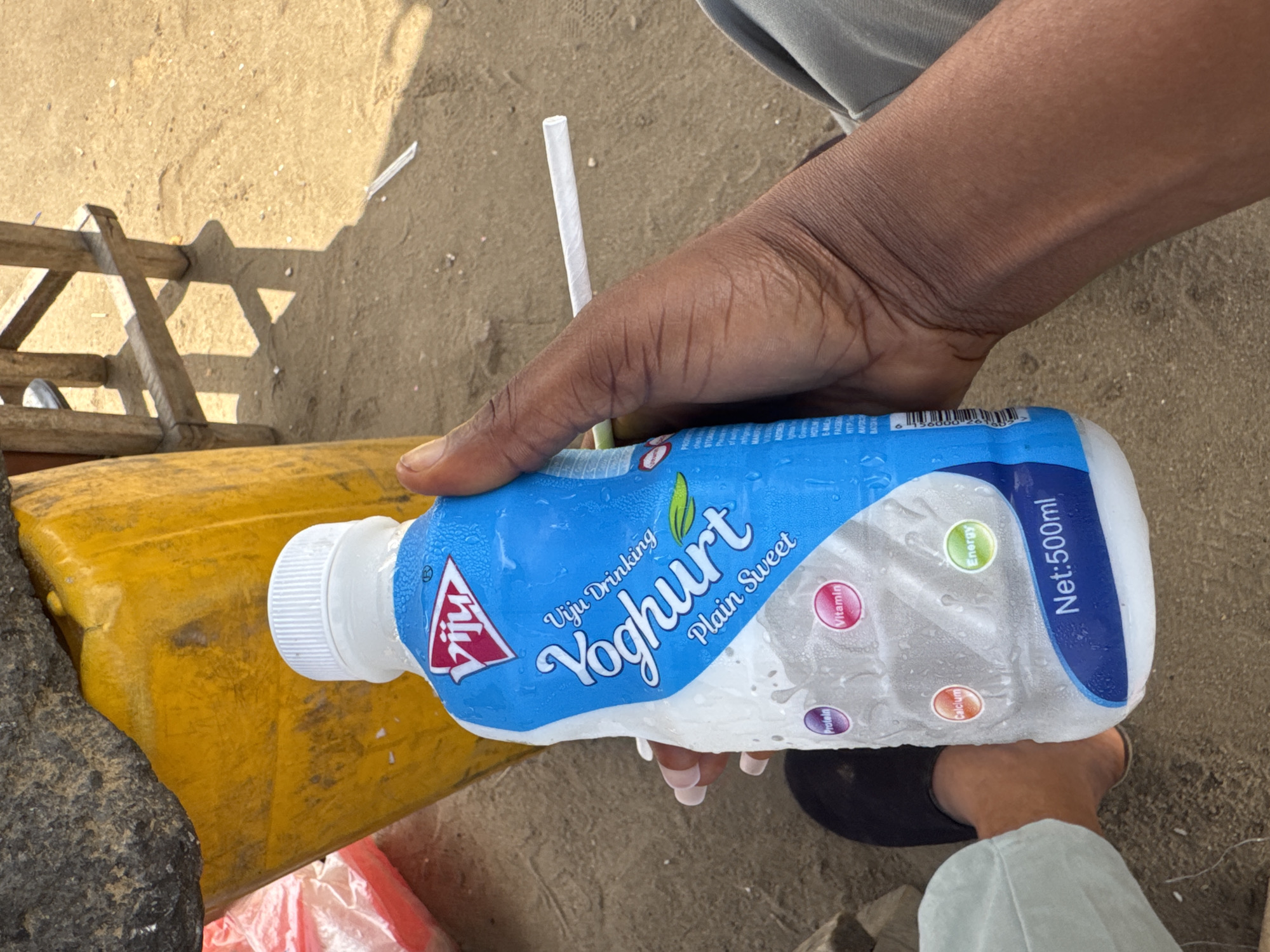 A dark-skinned hand holds a bottle of "Viju Yoghurt Plain Sweet" on a sunny, sandy surface. The bottle has condensation on its surface, indicating it is cold. A white drinking straw is inserted into the bottle. A yellow container is visible next to the bottle, along with a partial view of a wooden structure. The bottom portion of the image shows a foot in a black slipper. The brand name and descriptions on the bottle are legible, including nutrition information and the volume of 500ml.
The location in the city of Ajara, Nigeria cannot be determined from the image.
