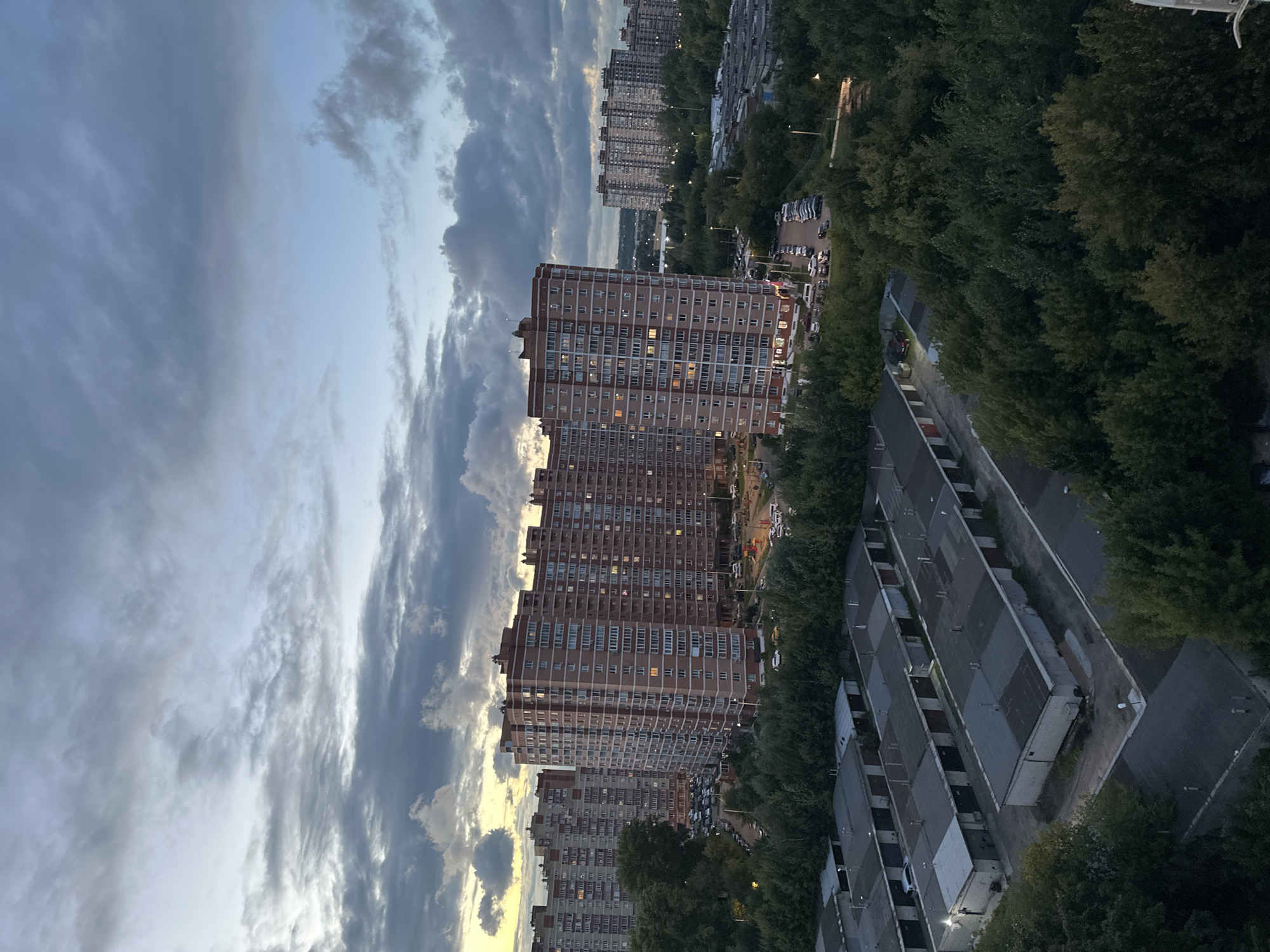 The image depicts an urban landscape in Shchyolkovo, Russia, at dusk. In the foreground, a row of identical, uniformly grey garages with darker grey roofs stretches across the frame. Beyond the garages, a dense canopy of dark green trees creates a natural barrier. In the mid-ground, multiple multi-story apartment buildings, constructed with reddish-brown bricks and featuring numerous windows, dominate the scene. A parking lot filled with various vehicles is visible in front of these buildings. Further in the distance, more apartment blocks are visible, interspersed with greenery and a body of water. The sky above is a blend of muted blues and greys, with dramatic clouds indicating an overcast sky, potentially before or after a rain shower. The overall impression is of a residential area in the late evening.