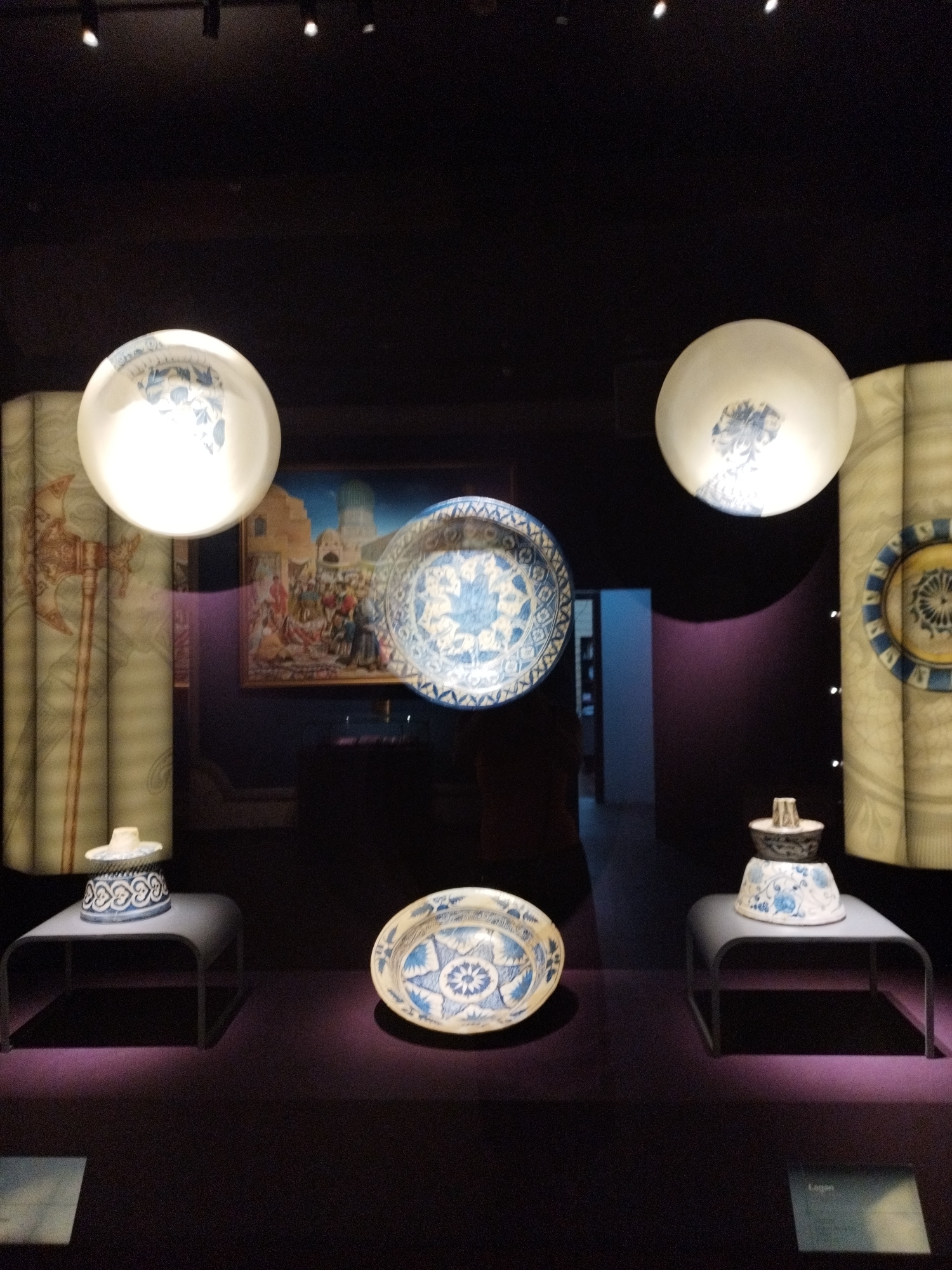 The image depicts an indoor exhibition space with museum-style displays. In the foreground, on a dark purple surface, are two blue and white ceramic objects. The object on the left is a shallow bowl with intricate floral patterns. To the right, a taller, lidded ceramic jar is displayed on a dark grey, curved shelf. Behind these, a large circular, white display object with blue floral patterns hangs from the ceiling, casting light. To the left and right of this hanging display are two tall, vertically oriented fabric panels with printed motifs, one showing an axe and the other a circular design. In the background, a framed painting or tapestry is visible, depicting a scene with numerous figures in a marketplace setting. Further back, a doorway or corridor is visible. The lighting is dim, with spotlights highlighting the displayed objects.