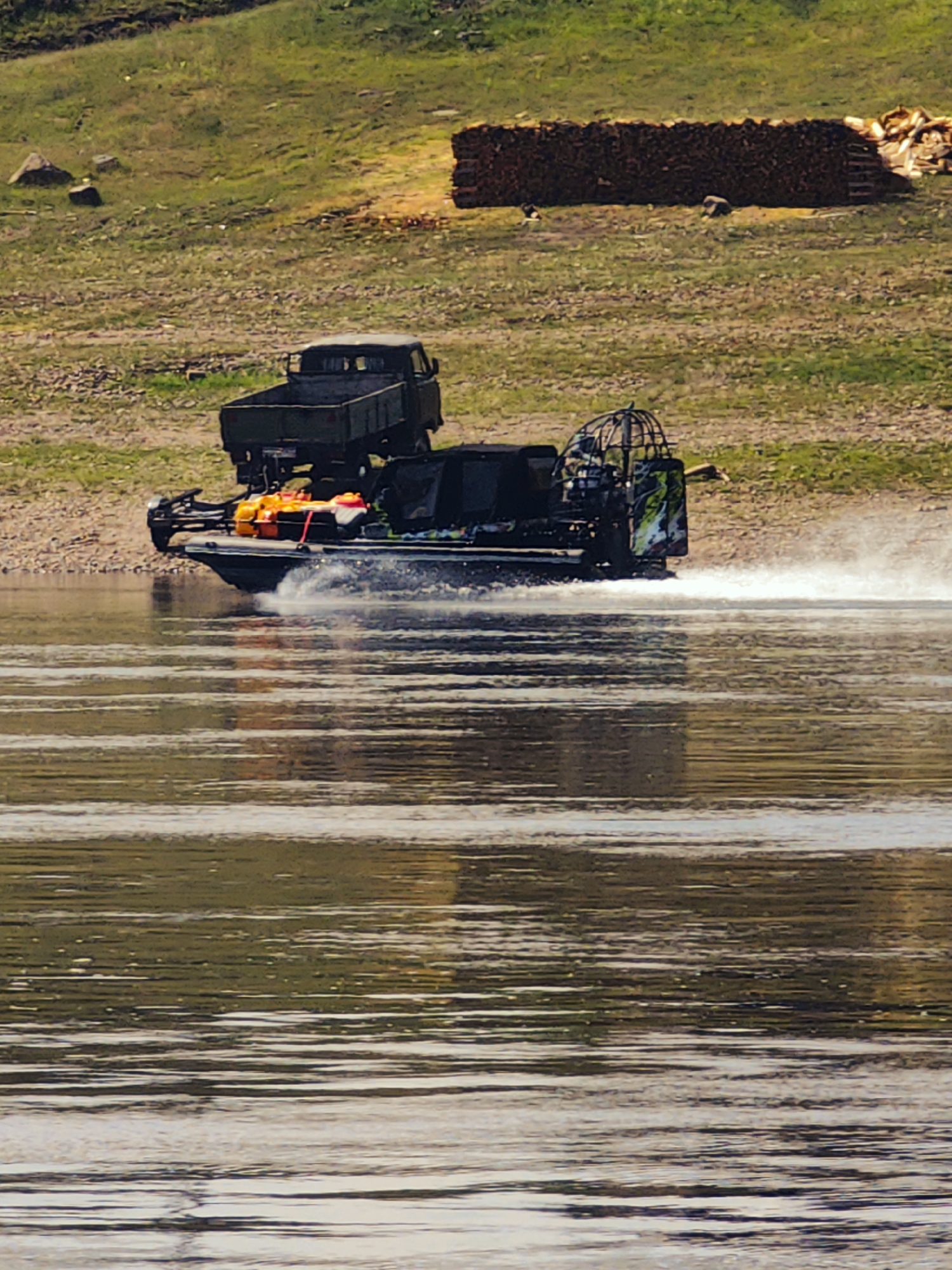 An amphibious vehicle, resembling a hovercraft with a truck bed, moves across a body of water. The vehicle is dark green and appears to be carrying cargo, including orange life vests, in its bed. The water is rippled, indicating movement. In the background, a grassy embankment slopes upwards, with scattered rocks and a large stack of wood visible. The overall scene suggests an outdoor, possibly rural or natural environment, with no immediate indication of a city or specific urban features. The location in Погодаево, Russia, is not visually discernible from the image itself.
