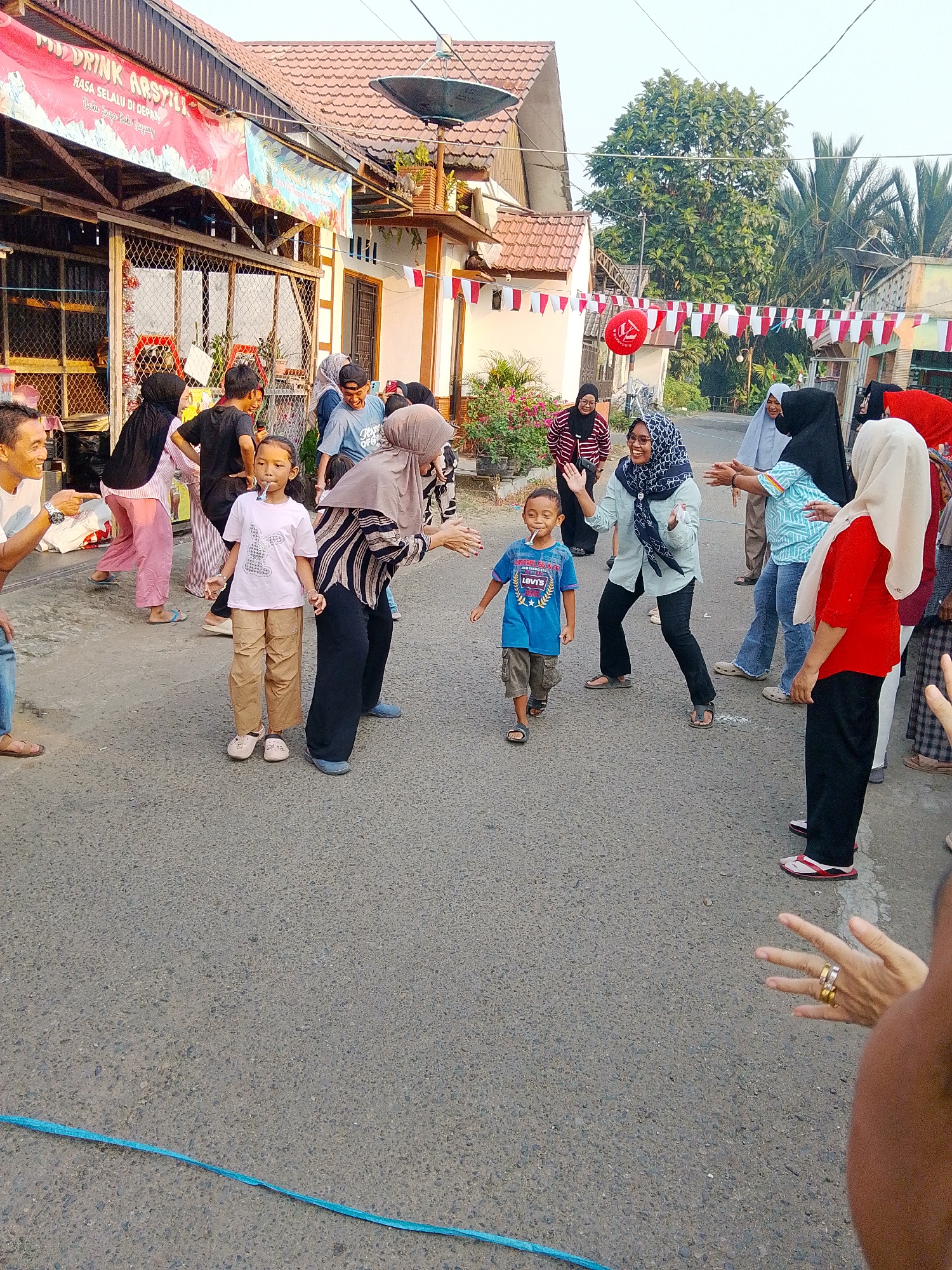 In Barabai, Indonesia, a group of people are gathered on a paved street, likely participating in a local celebration, given the Indonesian flags strung overhead. The scene is set in front of a building with a red-tiled roof and storefronts adorned with banners. In the foreground, a young boy in a blue t-shirt and shorts, holding a lollipop, walks towards the viewer while several women, some wearing hijabs, watch him. To the left, a young girl in a pink t-shirt and tan pants also holds a lollipop. A man in a white t-shirt points towards the left, and other individuals are visible in the background, engaged in conversation or observation. The atmosphere appears festive and communal.