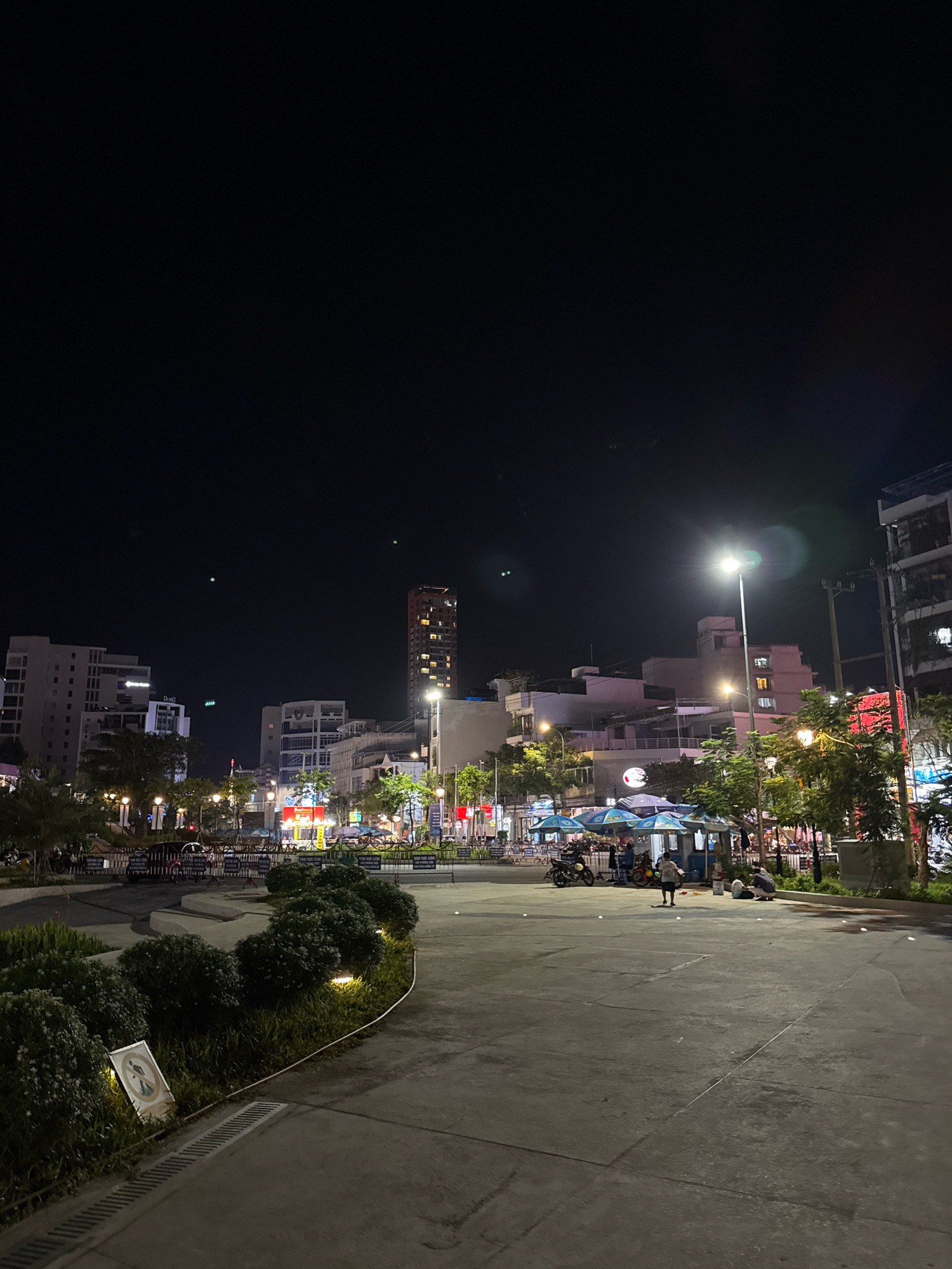 The image depicts a nighttime scene in Nha Trang, Vietnam, likely a public square or promenade adjacent to a street. Tall buildings with illuminated windows dominate the background, suggesting an urban environment. In the mid-ground, several street vendors' stalls are set up under blue and white umbrellas, with motorcycles parked nearby. People are visible at the stalls and sitting on the ground. The foreground features a paved walkway with manicured bushes lining its edge, illuminated by small spotlights. A sign with a graphic and Vietnamese text is propped up near the bushes. The overall atmosphere is lively, with artificial lights illuminating the area.