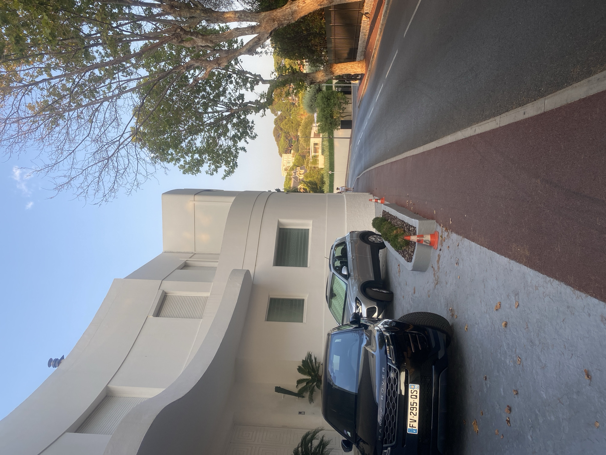 The image shows a modern white building with a curved facade, two parked cars, a street, and a sidewalk. The building has several windows with closed roll-down shutters. In the foreground, a black Range Rover is parked to the left, and a silver SUV is parked to its right. The cars are parked on a gray paved area. The street is paved and has white lane markings. A red and brown patterned sidewalk runs alongside the street. A person is walking on the sidewalk in the distance. Trees with green leaves line the street on the right side. The background shows a hillside with more buildings and trees, suggesting a suburban or semi-rural setting. The sky is clear and blue. The license plate on the black Range Rover reads "FV-295-QS".
Based on the presence of French license plates and the architectural style which is typical of the French Riviera, this image is likely located in the city of Antibes, France.