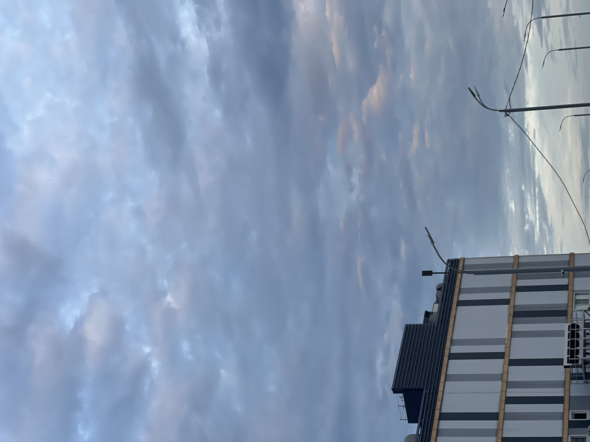 The image shows the upper portion of a modern building with a striped facade, featuring vertical stripes of white, gray, and dark gray, with a thin gold band separating two levels. The building has a dark gray, corrugated roofline. To the right of the building, a street lamp with a single light fixture is visible, along with a network of overhead wires. In the background, the sky is filled with a mix of gray and blue clouds, with some hints of pinkish light, suggesting either sunrise or sunset. The scene is likely located in an urban environment.