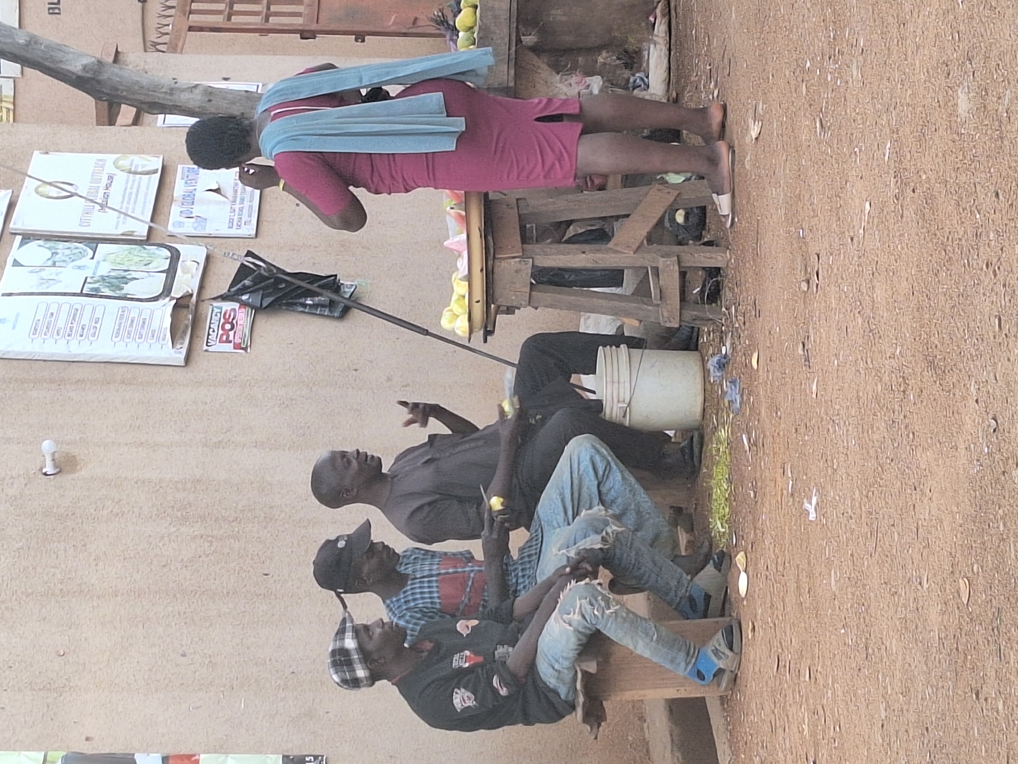 In Sabon Tasha, Nigeria, a street scene shows a woman in a rose-colored dress and light blue shawl standing at a wooden stall selling yellow fruits. Adjacent to her, three men are seated on a wooden bench along a wall. The man closest to the woman is wearing a dark gray shirt and black pants; the second is in a red and blue plaid shirt and ripped jeans; and the third is wearing a black T-shirt and ripped jeans. The men appear to be eating the same type of yellow fruit sold at the stall. Signage on the wall, including one indicating "VACANCY," is visible.