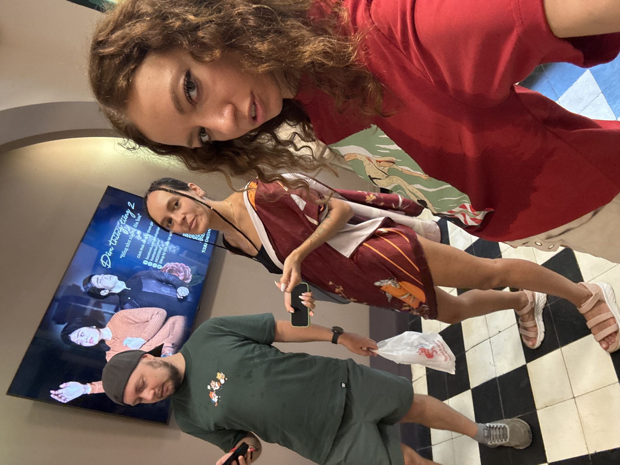 This indoor photograph shows three people on a black-and-white checkered floor. A woman with curly brown hair is in the upper right, wearing a red shirt with a graphic on the front. Below her is another woman with braided dark hair wearing a maroon dress with a white sash. She holds a smartphone. A man is below her, wearing a green T-shirt and shorts. He holds a smartphone in one hand and a white plastic bag in the other.
A TV screen is mounted on the wall behind them, displaying images of a couple in an advertisement. Text in Vietnamese is also visible on the screen, likely indicating the advertisement is related to a local business. The text includes the phrase "Đêm thiêng liêng 2" and mentions the city of Hà Nội, Vietnam, suggesting the location of the photo.