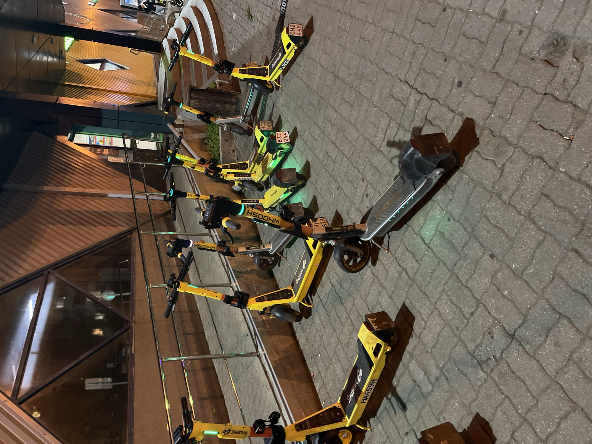 A row of yellow electric scooters is parked on a paved walkway in Mytishchi, Russia, in front of a building with a modern, angular facade. The scooters, some branded "WHOOSH" and "Яндекс Go," have their kickstands down. Several of the scooters display license plates, including "HA459H," "HY 87A," "HC8 360," "HC8 244," and "OM2 44Y." The ground is paved with interlocking bricks, and a metal railing runs along the walkway. In the background, the building's facade features patterned metal panels and large windows that reflect ambient light. The scene is illuminated by artificial lighting, suggesting it is nighttime or dusk. No people are clearly visible interacting with the scooters.