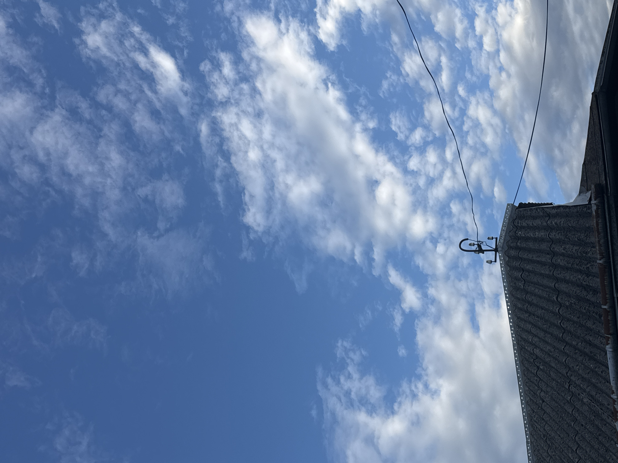 The image shows a close-up, low-angle view of a dark, corrugated rooftop against a bright blue sky with scattered white clouds. The rooftop is textured and appears weathered. At the peak of the roof, there is a dark metal fixture with a curved top and two smaller horizontal arms extending outwards. Two dark electrical wires run horizontally across the frame, one above the other, against the backdrop of the sky and clouds. The scene is devoid of people and any explicit indication of a specific city or location. The overall impression is one of an overcast day with a focus on the sky and man-made elements on the roof.