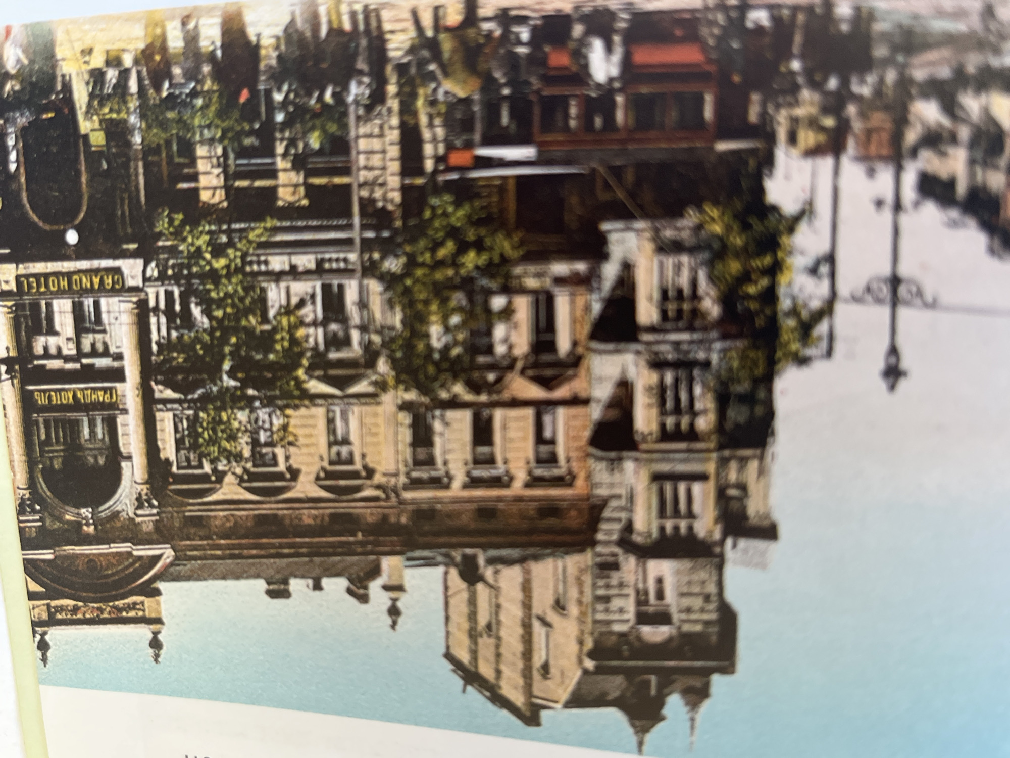 The image depicts a sepia-toned postcard showing a grand hotel facade with ornate architectural details. The hotel has multiple stories with numerous windows and balconies. A sign on the building reads "GRAND HOTEL" in yellow lettering. In front of the hotel, there appears to be a street with some greenery and possibly a tram or trolley. The overall impression is of a historic European city.