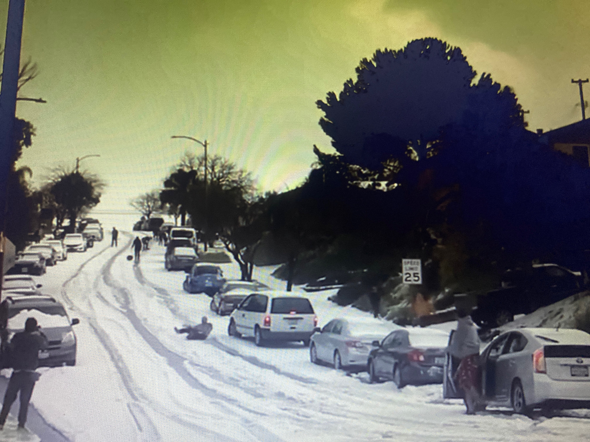 The image depicts a snowy street scene in Hemayetpur, Bangladesh, with several cars parked along the sides of the road. A few people are visible, some walking and others seemingly engaged in recreational activities on the snow-covered ground. One individual appears to be sledding down the street. The sky is a hazy yellow-green. There is a speed limit sign showing "25".