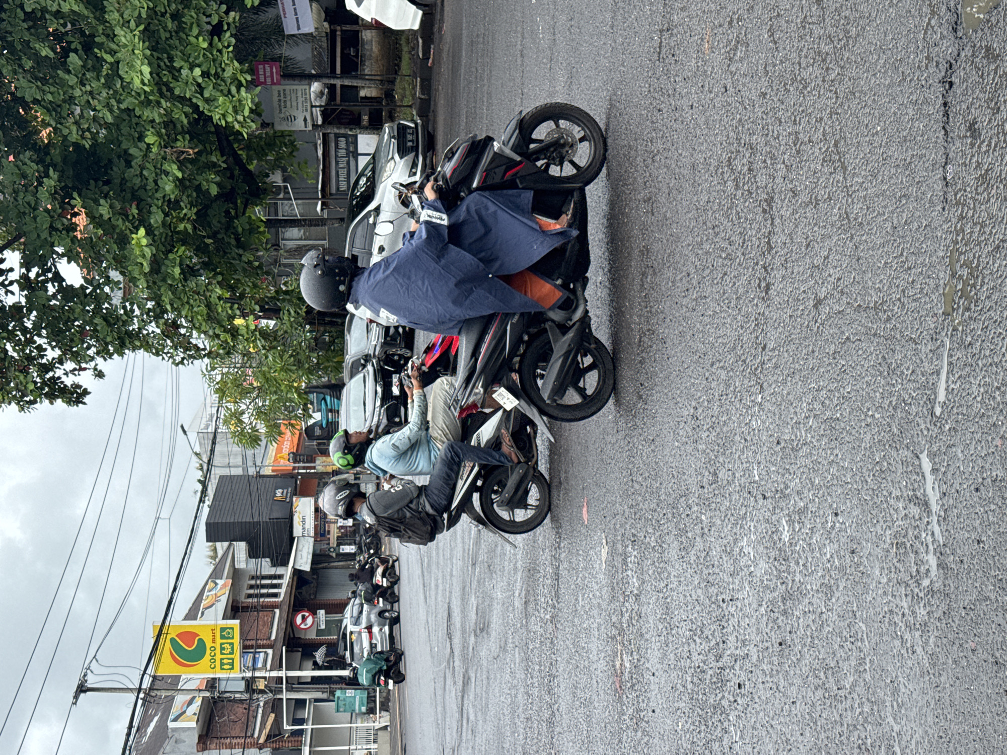 This image depicts a bustling street scene in Kuta Selatan, Indonesia, under overcast skies, with the wet asphalt road indicating recent rain. The central focus is a line of people on motorcycles and scooters, patiently waiting, suggesting traffic or a temporary stop.
In the mid-ground, a person wearing a black helmet and a dark blue rain poncho over orange trousers is seated on a black and red scooter. Further down the road, other riders are visible, also wearing helmets. One rider wears a light blue long-sleeved shirt and beige pants on a white scooter, while another wears a dark backpack and dark attire on a black scooter. A motorcycle with a green backpack is also partially visible on the left.
The left side of the street is lined with lush green trees, while the right side features various commercial buildings. Several signs are visible, providing clear location cues: "COCO mart" (a local convenience store chain), "NASI PECEL MAS TIO SOLO" (a restaurant), "mandiri" (an Indonesian bank), and signs for "bumble bee kids zone" and "KIDS THERAPY." A partial sign reads "PERHAT," likely short for "PERHATIAN" (Attention/Caution). Utility poles with numerous overhead wires are also prominent. The scene captures a typical urban environment in Southeast Asia, with motorcycles as a primary mode of transportation, adapting to the local weather conditions.