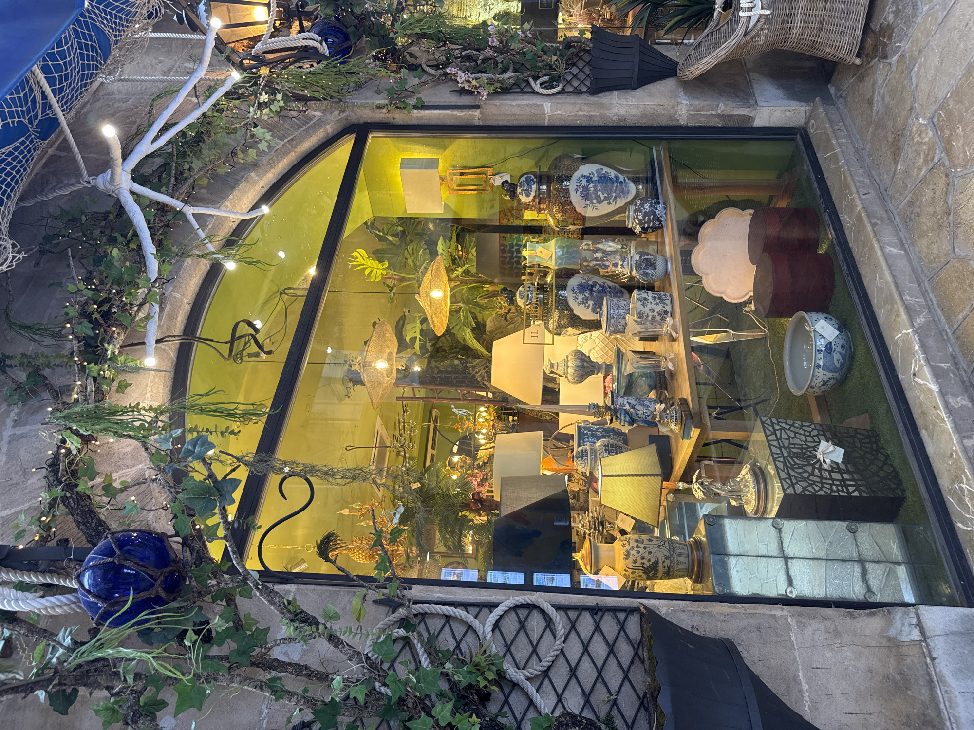 The image shows a store window display on a street in Palma de Mallorca, Spain. The window features a collection of decorative items, including lamps, vases, and decorative bowls, many of which are in blue and white porcelain. Green foliage and artificial plants are integrated into the display, along with string lights and ropes, creating a somewhat whimsical and nautical theme. The window itself is arched and framed by stone. Inside the store, more decorative items and lighting fixtures are visible. The exterior of the store appears to be constructed of stone, and the street in front is paved with irregular stones. There are no people visible in the image.