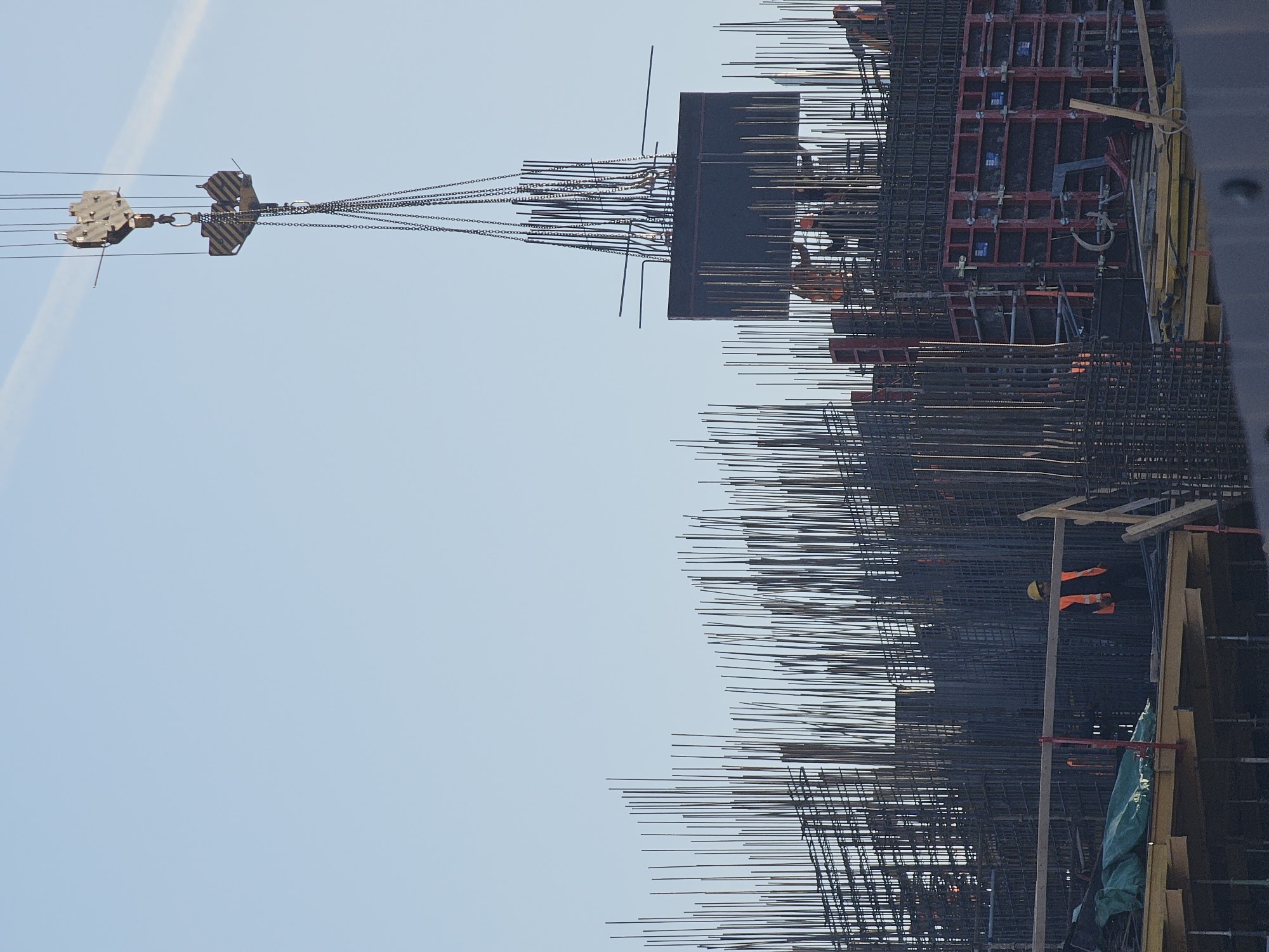 The image shows a construction site in Moscow, Russia. A crane is lifting a section of rebar and formwork for a large concrete structure. Workers in safety gear are visible amongst the rebar. The sky is pale blue.