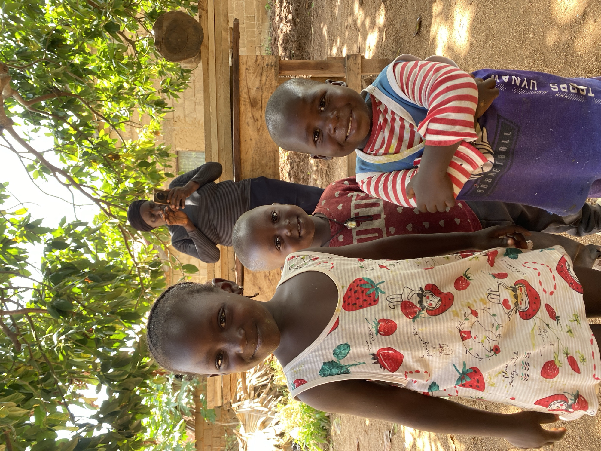 A person is taking a photograph of three children outside, near a building. The child in the front is wearing a white dress with strawberry designs, the middle child is wearing a red patterned shirt, and the child in the back is wearing a striped shirt and is wrapped in a blue cloth. The photo was taken during the day. This image shows a rural landscape, which could be the city of Riyom, Nigeria.