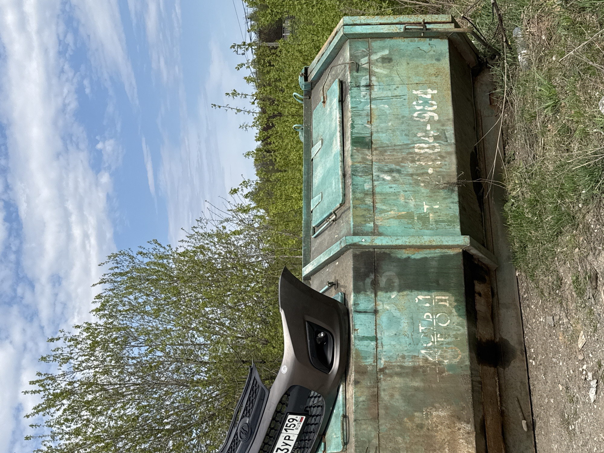 The image depicts a rusted, green metal utility box in an outdoor setting. The box has white painted numbers "736-78-984" and Cyrillic script on its side. Leaning against it is a gray bumper and part of the grill of a vehicle with a Russian license plate that reads "3YP 159 RUS". The surrounding environment includes trees, grass, and a cloudy sky. Based on the license plate code "159", the location is identified as the city of Perm, Russia.