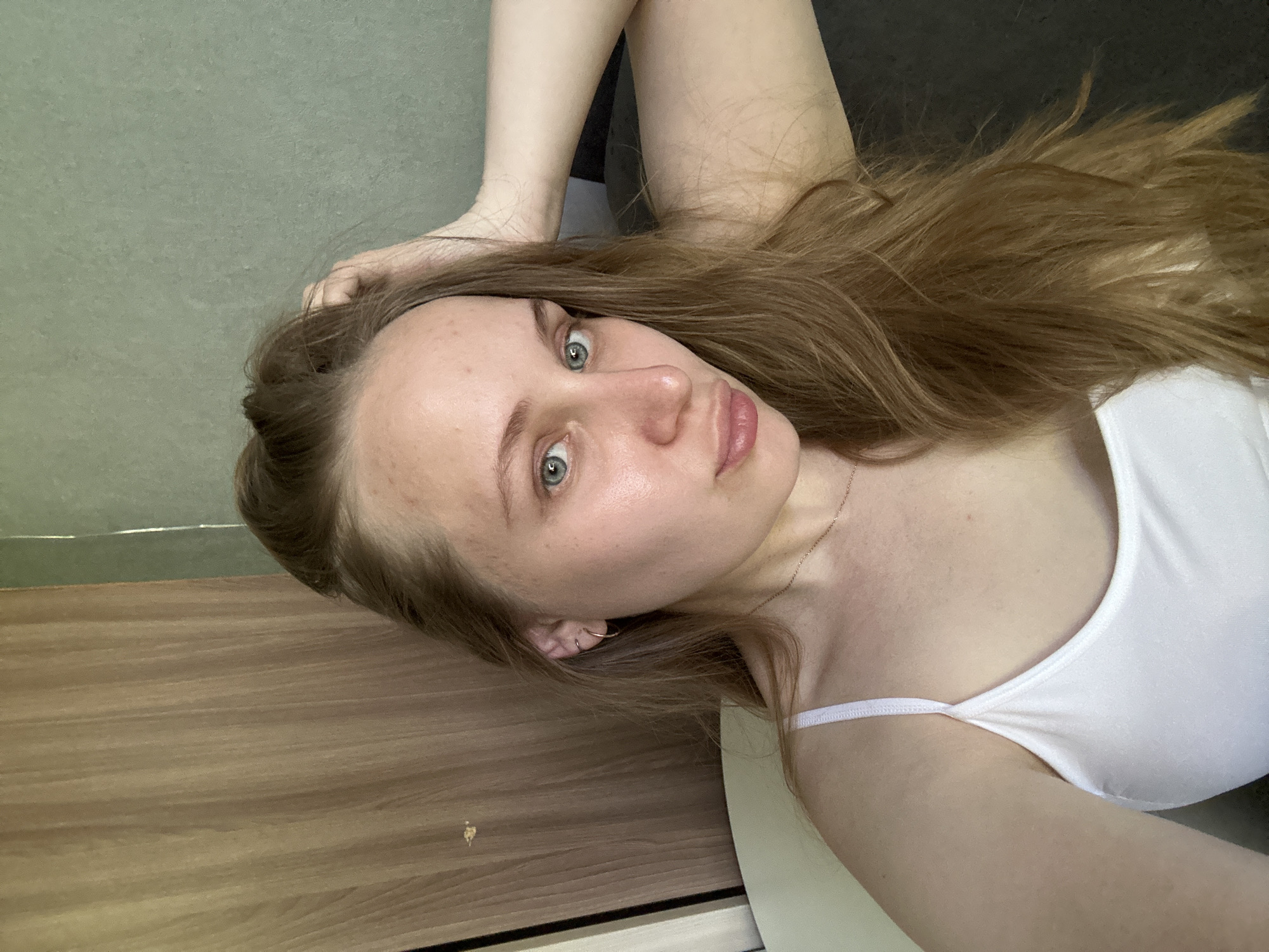 A young woman with long, light brown hair and blue eyes is pictured indoors. She is wearing a white tank top. The background is a combination of a light brown wood-grain surface and a gray wall. The photo appears to be taken at an angle. Location is not identifiable as Chaikovsky, Russia.