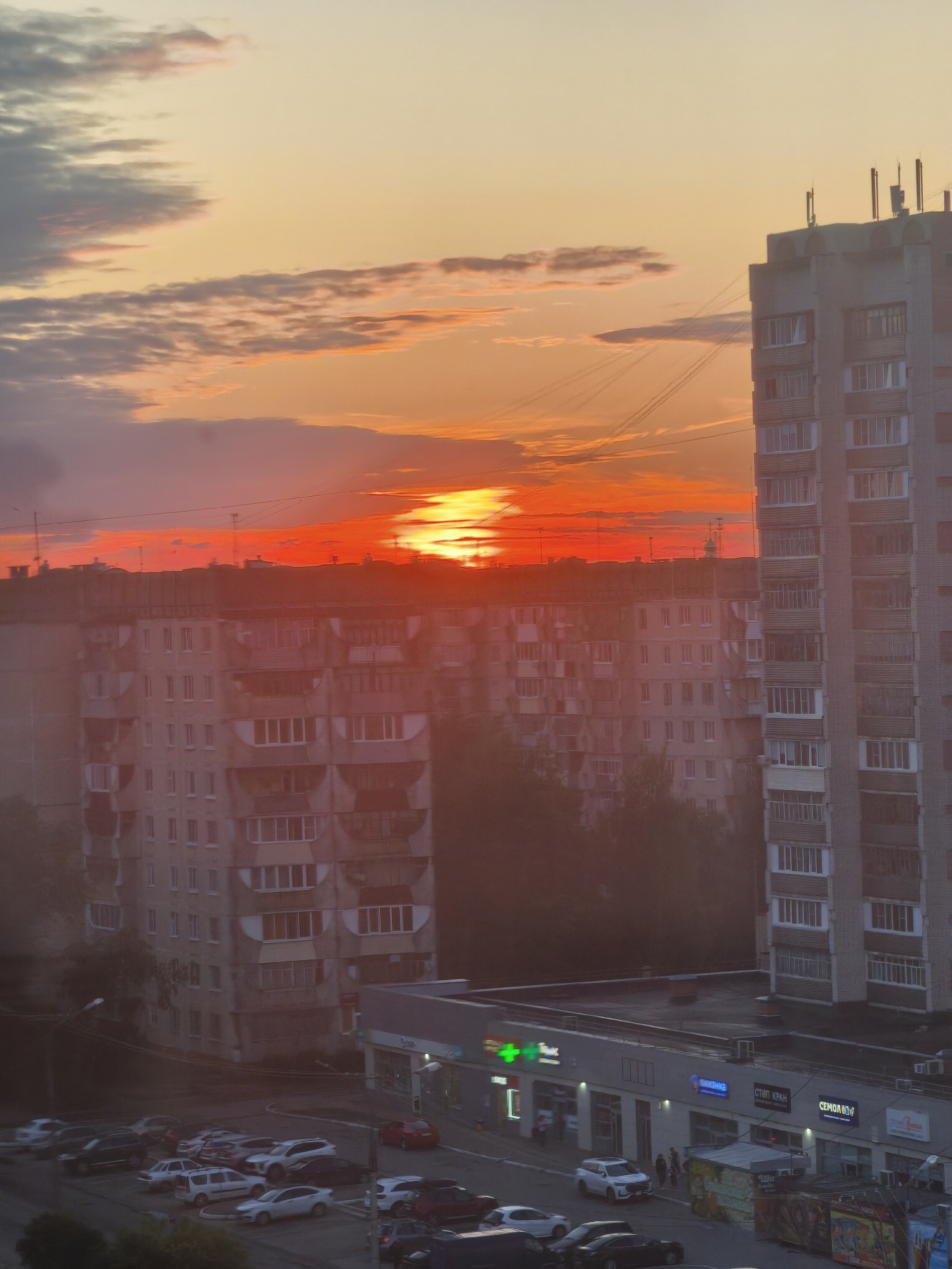 The image captures a sunset over a residential area in Yoshkar-Ola, Russia. The sky is filled with vibrant hues of orange and red, indicating the sun is setting or has just set. Several tall residential buildings, characteristic of Soviet-era architecture, dominate the skyline. The building on the right is a light-colored brick structure with multiple windows. The building in the middle ground appears to be older and is painted in a lighter color, with balconies visible on some floors. Below these buildings, a street level view reveals a parking lot filled with various cars. A few people are walking on the sidewalk in front of a row of shops. The shops have illuminated signage, with one sign appearing to be in Russian. The overall impression is a glimpse into urban life in Yoshkar-Ola during twilight.