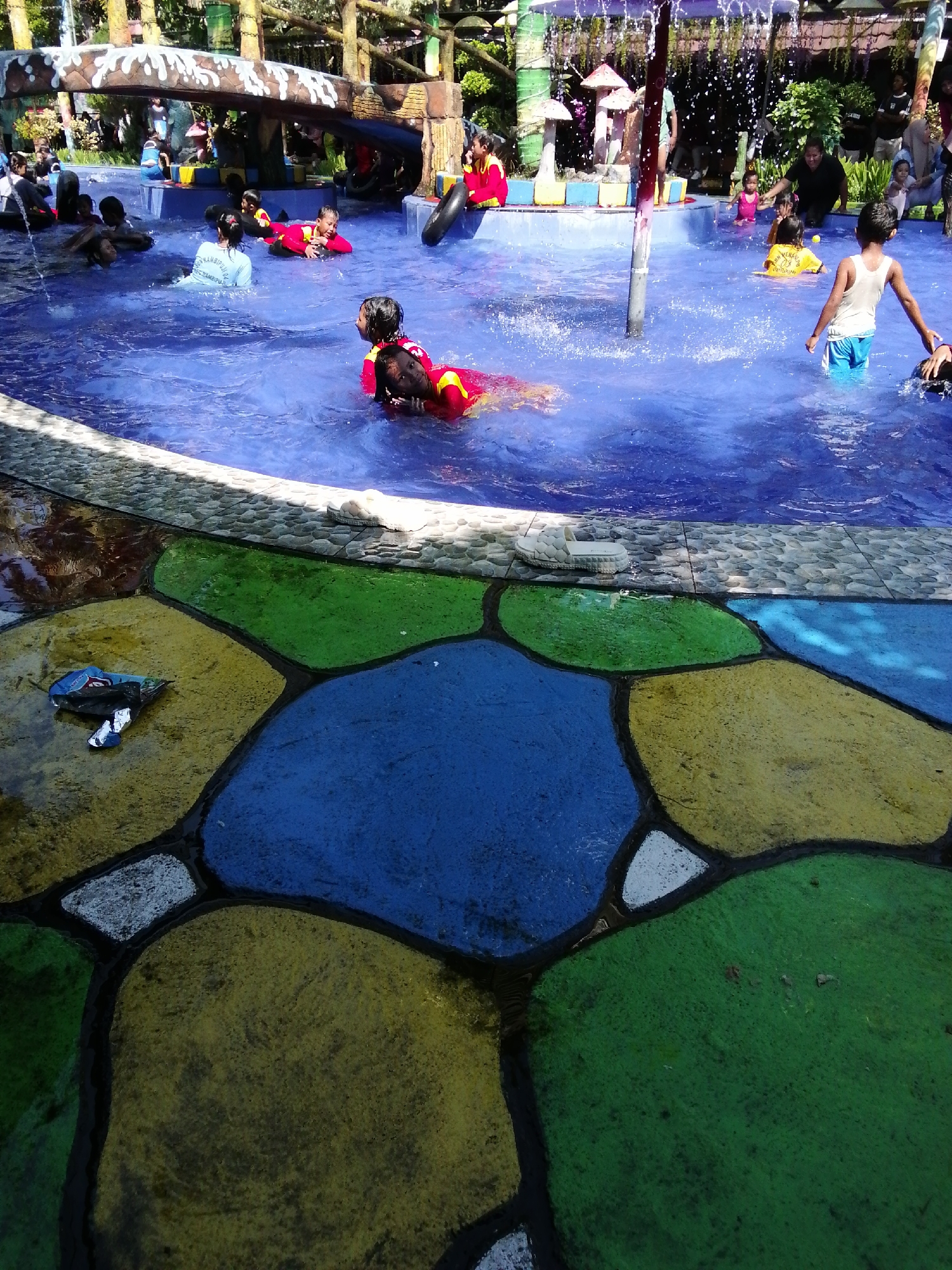 The image depicts a busy outdoor swimming pool, likely part of a water park, situated in Tegalwangi, Indonesia. The pool is filled with many children and a few adults engaged in various water activities. Several children are in the water, some wearing red and yellow life vests, others in casual swimwear. One child, seen from the back, is wearing a white tank top and blue shorts. The pool's edge is decorated with large, brightly colored, irregular-shaped tiles in blue, green, and yellow. In the foreground, on the tiled area next to the pool, there is a discarded plastic wrapper and a white shoe. In the background, there is a jungle-gym-like structure with slides and platforms, and numerous trees and foliage suggesting a tropical environment. The overall atmosphere appears to be one of recreation and family enjoyment.