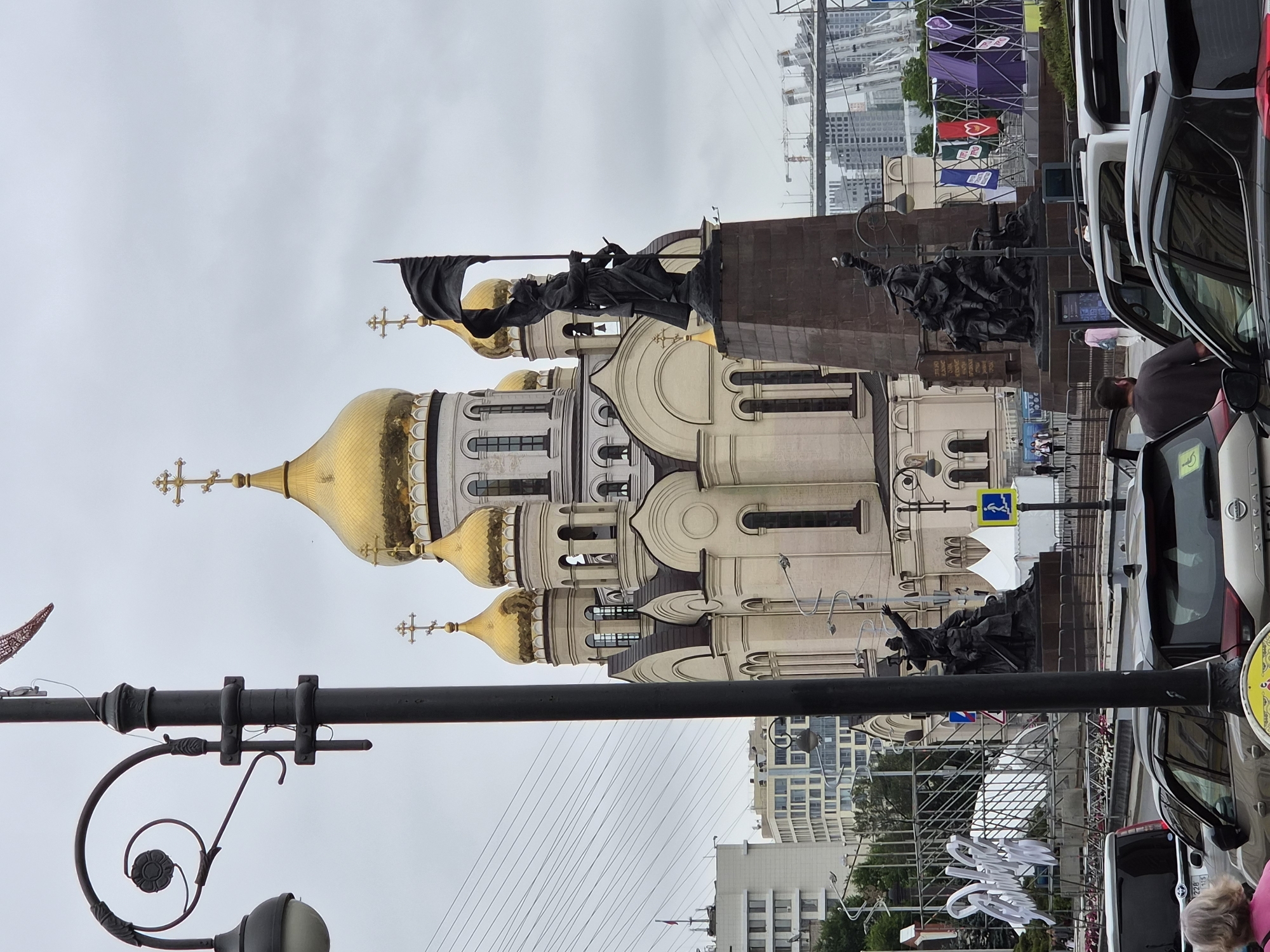 The image shows a view of the Svetlanskaya Street area in Vladivostok, Russia, looking towards the Church of the Archangel Michael. In the foreground, a black lamppost with an ornate scroll design is positioned off-center to the left. Behind it, the prominent golden domes of the church rise against an overcast sky. To the right of the church, a bronze monument stands on a pedestal, featuring several figures, including one holding a flag. The base of the monument has inscriptions. In the middle ground, there are various structures including a white tent and a large metal frame with white lettering. Further back, modern buildings and construction cranes are visible, suggesting ongoing development in the city. Several cars are parked or driving on the street in the foreground and middle ground, and a few pedestrians can be seen walking. The overall scene depicts an urban environment with a mix of historical and modern architecture, under a cloudy sky.