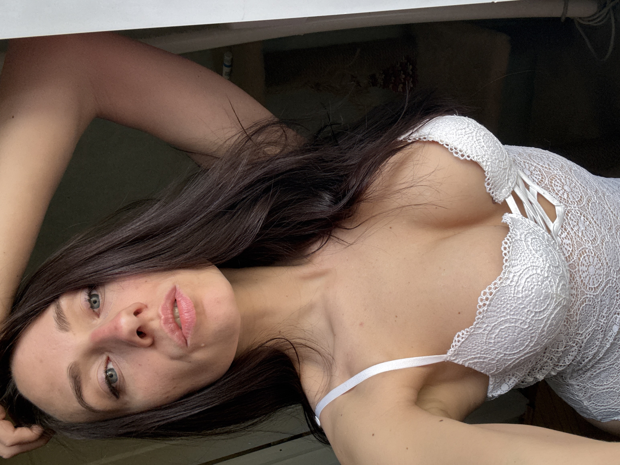 The image displays a woman posing indoors in Saint Petersburg, Russia. She is shown in a reclined or lying position, looking directly at the viewer with a direct and focused gaze. Her head is slightly tilted, and her long, dark hair frames her shoulders and chest. She possesses fair skin, light-colored eyes, and full lips with light pink lipstick. A small mole is visible on her upper left arm. The woman is wearing a white lace lingerie piece or bodysuit, featuring intricate floral lace patterns and delicate strap details around the bust. The background is dark and indistinct, suggesting an interior setting, with a white ledge or window frame visible along the top and left edges of the frame. Soft, even lighting illuminates her features and the texture of her garment. There is no visible text in the image.