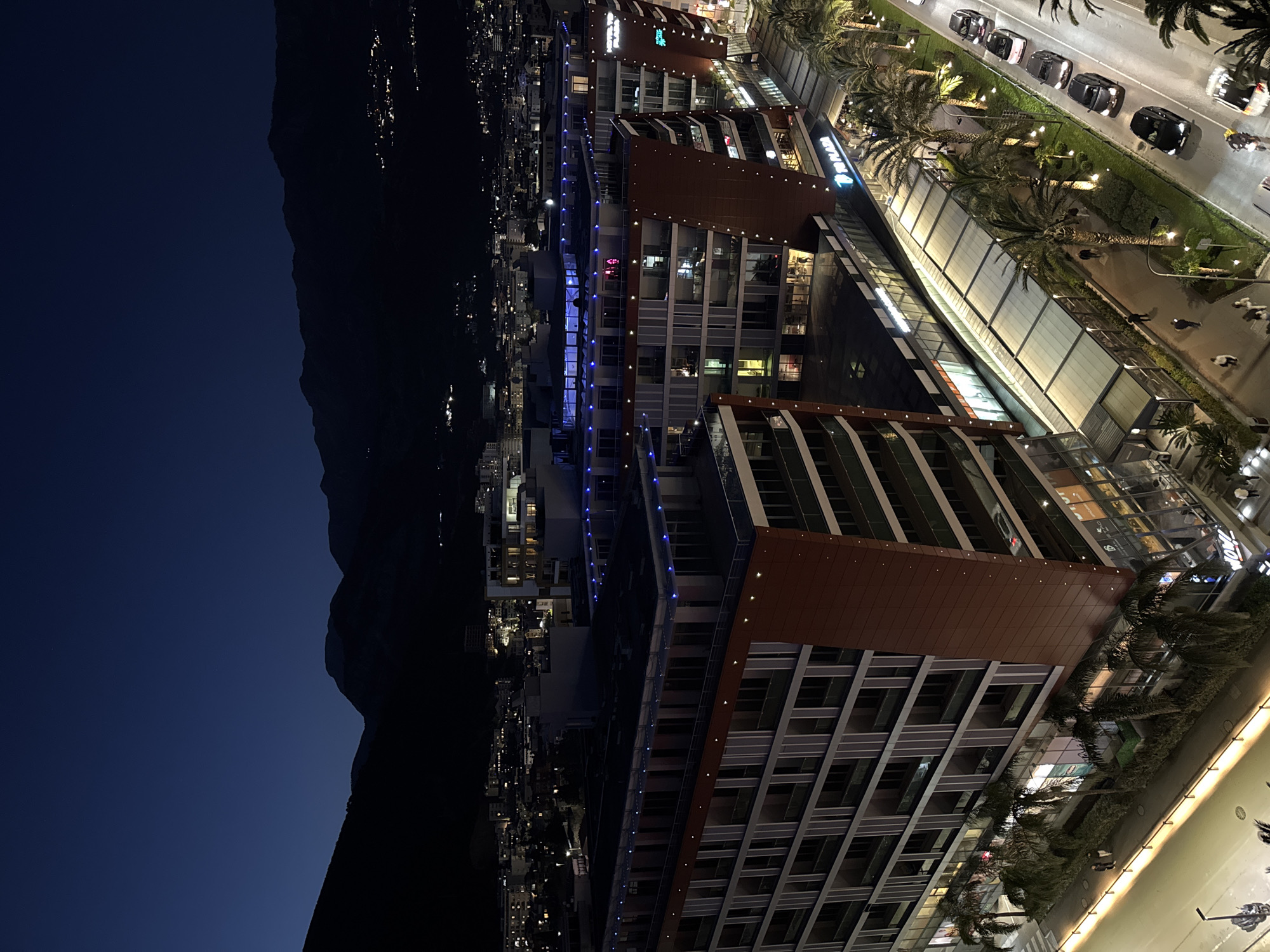 The image depicts a nighttime cityscape in Budva, Montenegro, taken from a high vantage point. In the left portion of the frame, a dark mountain range is visible against a deep blue twilight sky. To the right, illuminated buildings dominate the scene. These modern structures feature a mix of residential and commercial spaces, characterized by large windows and distinct architectural designs.
Between the buildings, the city lights twinkle, marking a busy urban environment. Along the right edge, a well-lit street scene unfolds, showing cars moving along the road and pedestrians strolling along the sidewalks shaded by palm trees. The atmosphere suggests a vibrant and bustling nightlife.