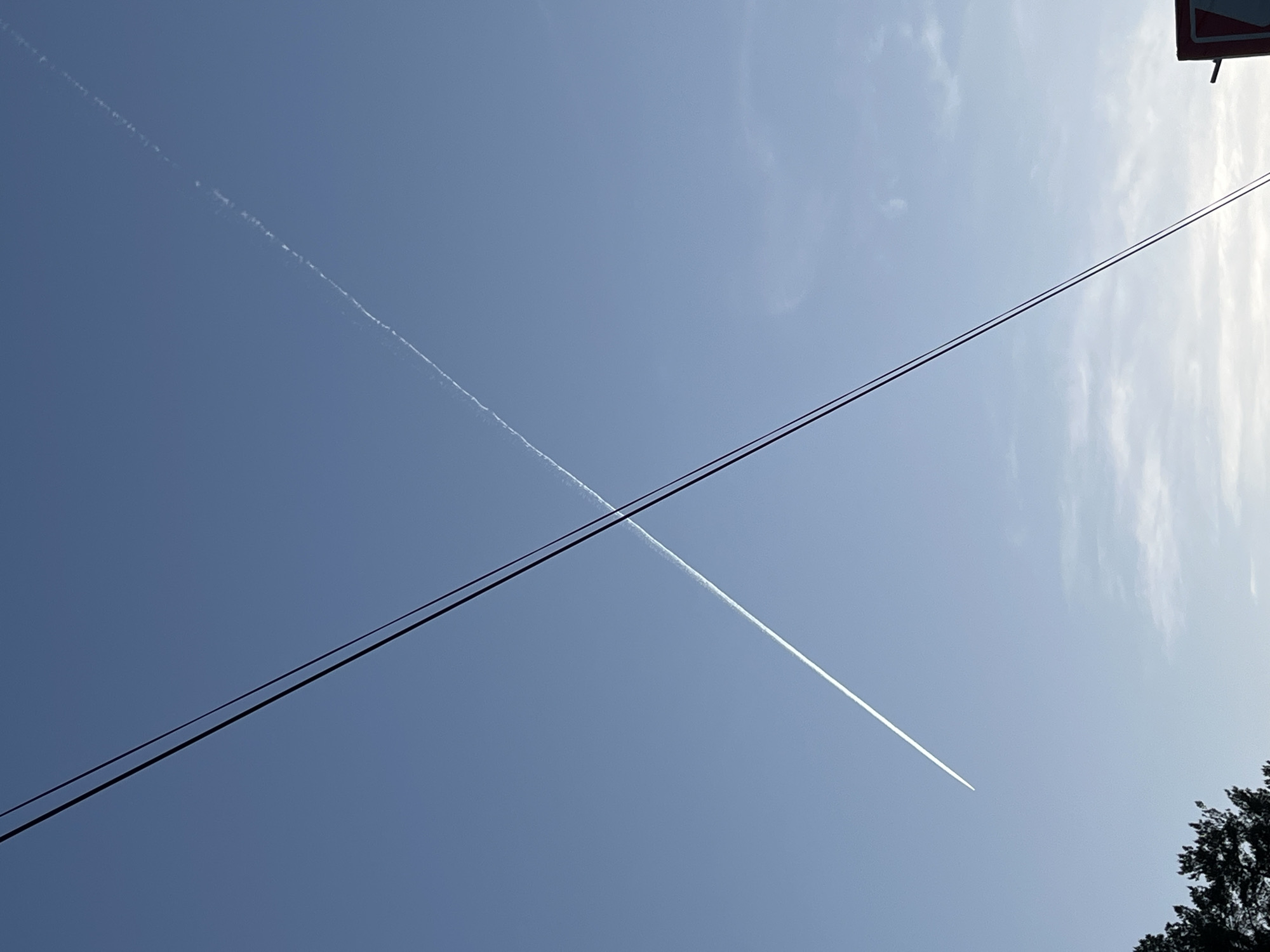 The image depicts a blue sky with two intersecting contrails. Two black power lines stretch diagonally across the frame from the upper right to the lower left. Some tree foliage is visible in the bottom right corner, and a blurred sign is in the upper right. The contrails create an X shape in the sky. The location is difficult to determine as the image only shows the sky. There is no indication of it being the city of Chaikovsky, Russia.