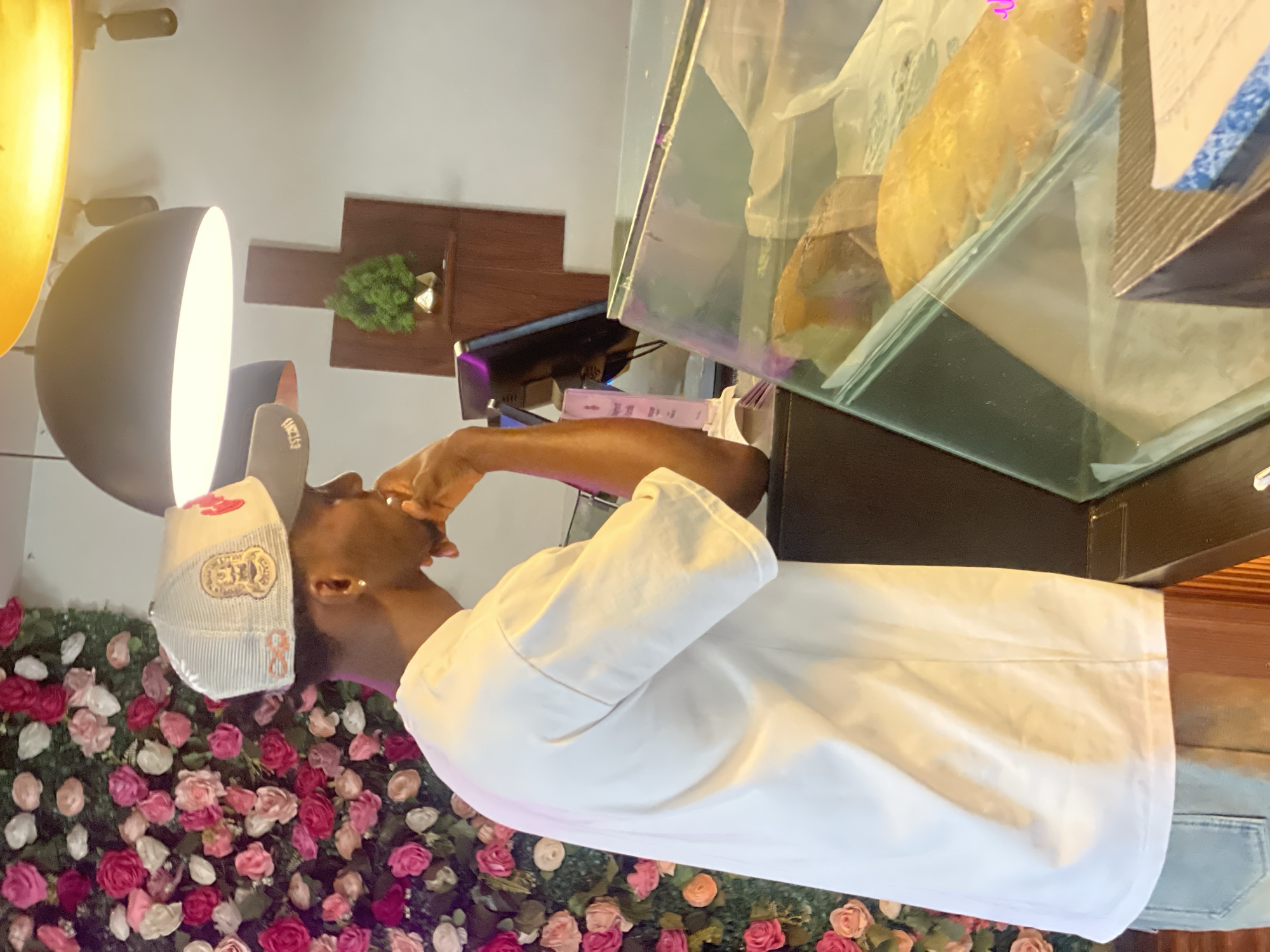 A person wearing a cap and a white shirt stands near a display case containing wrapped bread, possibly in a bakery or food establishment. A wall of pink and red roses is visible behind the person. Additional background elements include a lamp and some wooden decorative pieces on the wall. The location may be Adankolo, Nigeria.