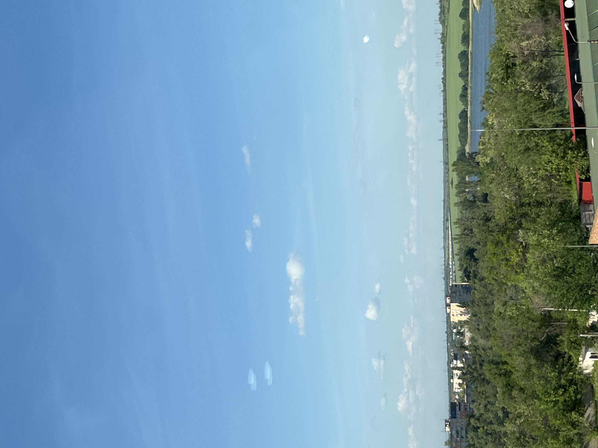 An elevated view shows part of Kryvyi Rih, Ukraine. The scene consists of a bright blue sky with wisps of clouds, the edge of a cityscape with buildings, trees, and greenery. A body of water is visible in the distance. A bit of a structure appears on the right, possibly part of a building.