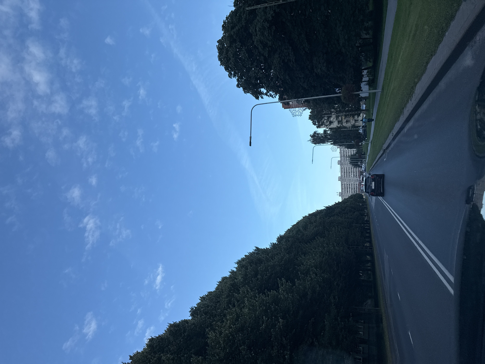 The image shows a view from a vehicle driving on a paved road in Tallinn, Estonia. The road is lined with trees on the left and a grassy median and sidewalk on the right. In the distance, there are several multi-story buildings. A black Mini Cooper is visible driving ahead on the road. The sky is blue with scattered white clouds. The time of day appears to be late afternoon or early evening.