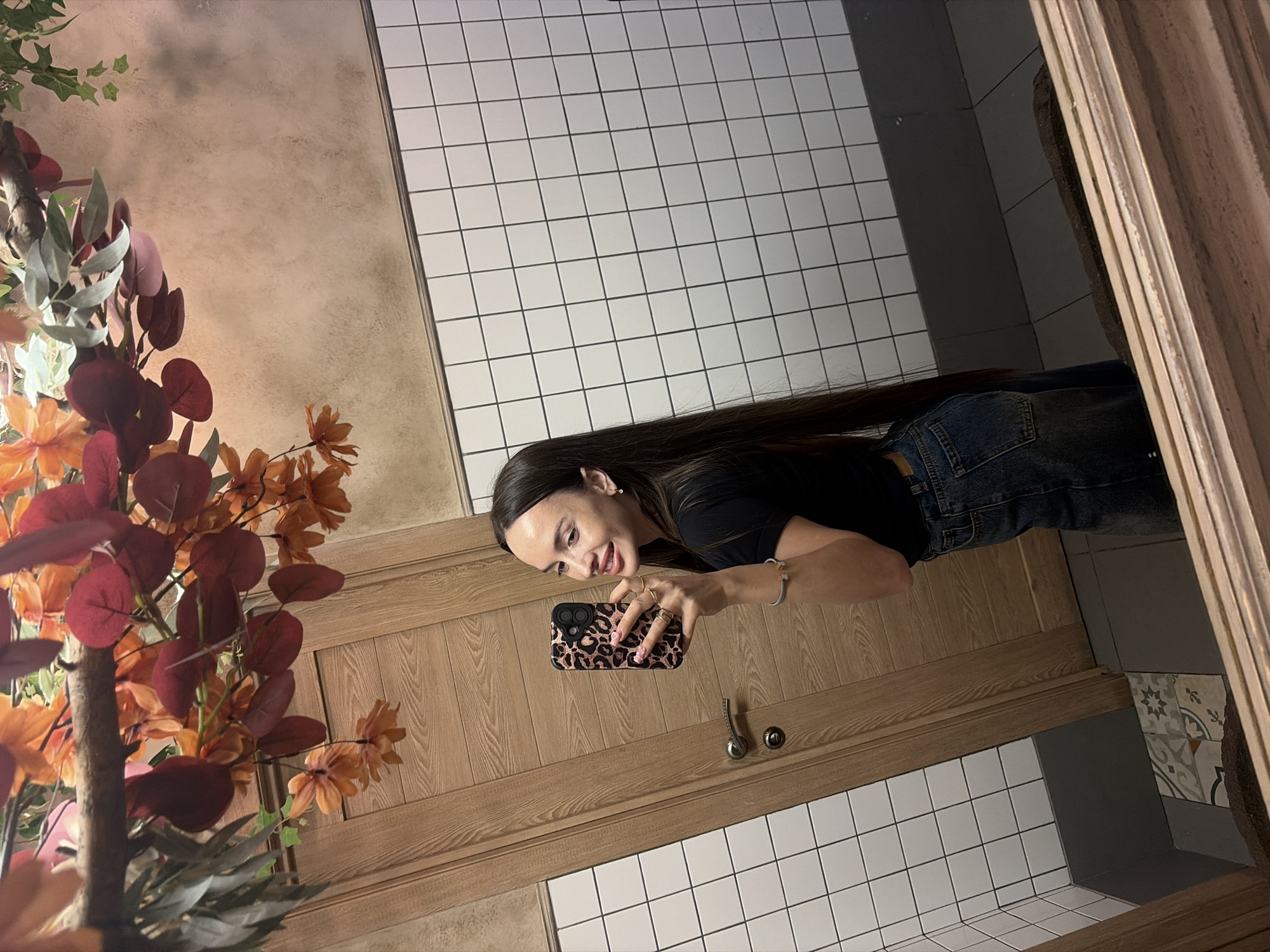 A young woman with long, dark hair is depicted taking a mirror selfie in an indoor setting, likely a restroom or a vanity area, possibly in Izhevsk, Russia. She is smiling and looking towards the camera, holding a smartphone with a distinct leopard print case in her right hand. She is wearing a black t-shirt, blue jeans, a bracelet on her left wrist, and rings on her right hand.
The background features a light wood-grained counter or vanity with two silver-colored knobs. Above this counter, to the left, is a vibrant decorative arrangement of artificial foliage and flowers, predominantly in shades of deep red, orange, and green. The walls around the woman and above the counter are covered with white square tiles with dark grout lines. In the upper left portion of the image (when oriented upright), a textured, light brown or beige wall surface is visible. To the bottom right, beyond the tiled area, there are patterned tiles with geometric and floral designs in shades of grey and white. The lighting appears to be artificial and internal to the building. There is no visible text in the image.