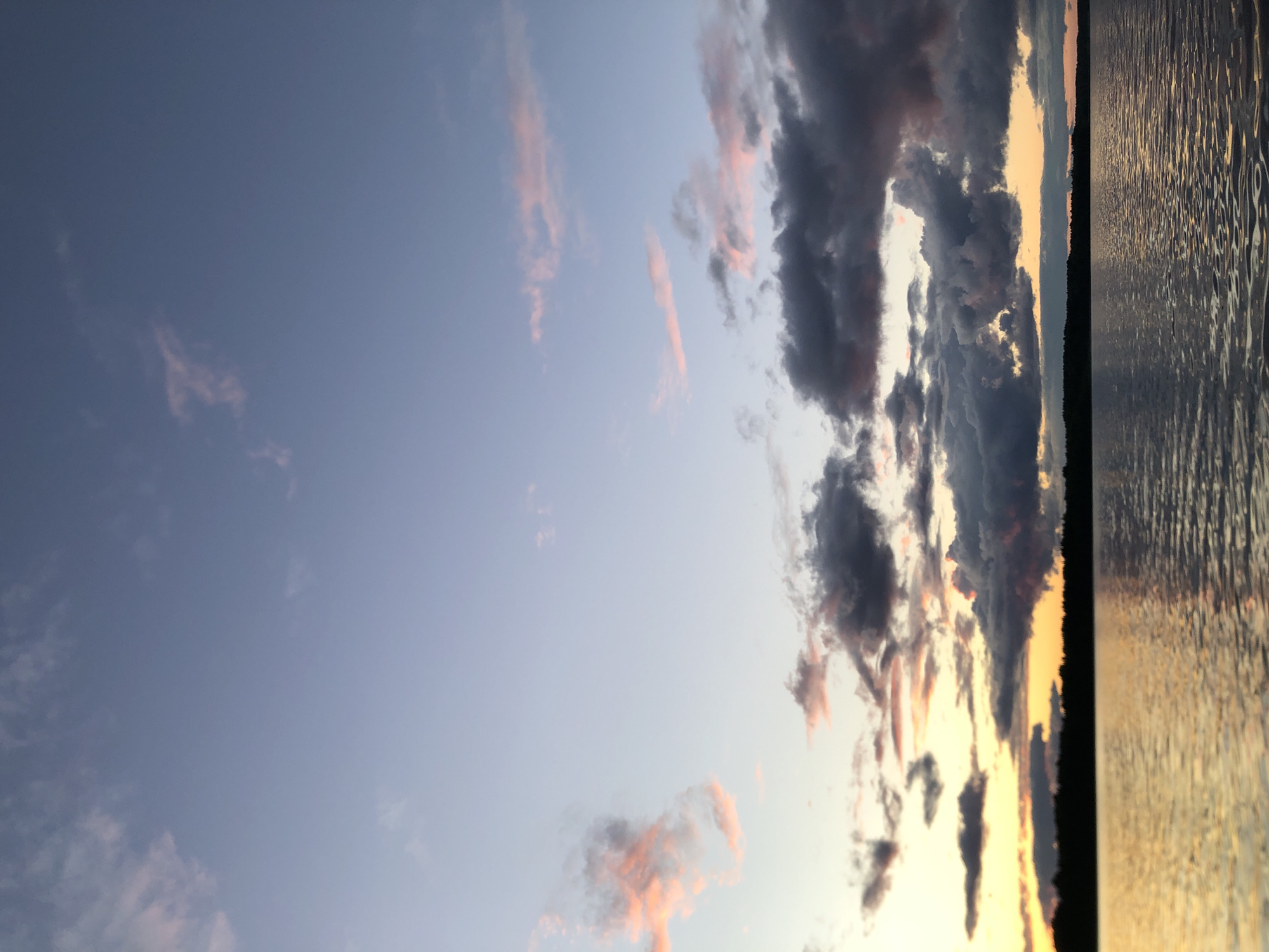 The media file captures a serene natural scene at dusk, likely in Svirstroy, Russia. The composition is divided vertically, with the left two-thirds dominated by a vast expanse of sky and the right third by a body of water.
The sky transitions from a soft, pale blue at the top to warmer hues of orange and yellow near the horizon, indicative of sunset. Wispy clouds are scattered throughout the sky; some are tinged with pink from the setting sun, while others are darker gray, creating a dramatic contrast.
The right side of the image features a tranquil body of water, possibly a lake or river, reflecting the colors of the sky. The water's surface is textured with ripples, catching the light and creating a shimmering effect. A dark, tree-lined horizon separates the sky from the water, suggesting a natural landscape.
The overall mood is peaceful and contemplative, evoking the beauty of a natural sunset. The time of day is clearly evening, as indicated by the warm light and cloud colors. There is no visible text in the image. The scene appears to be an outdoor, natural setting, with no people or man-made objects prominently featured.