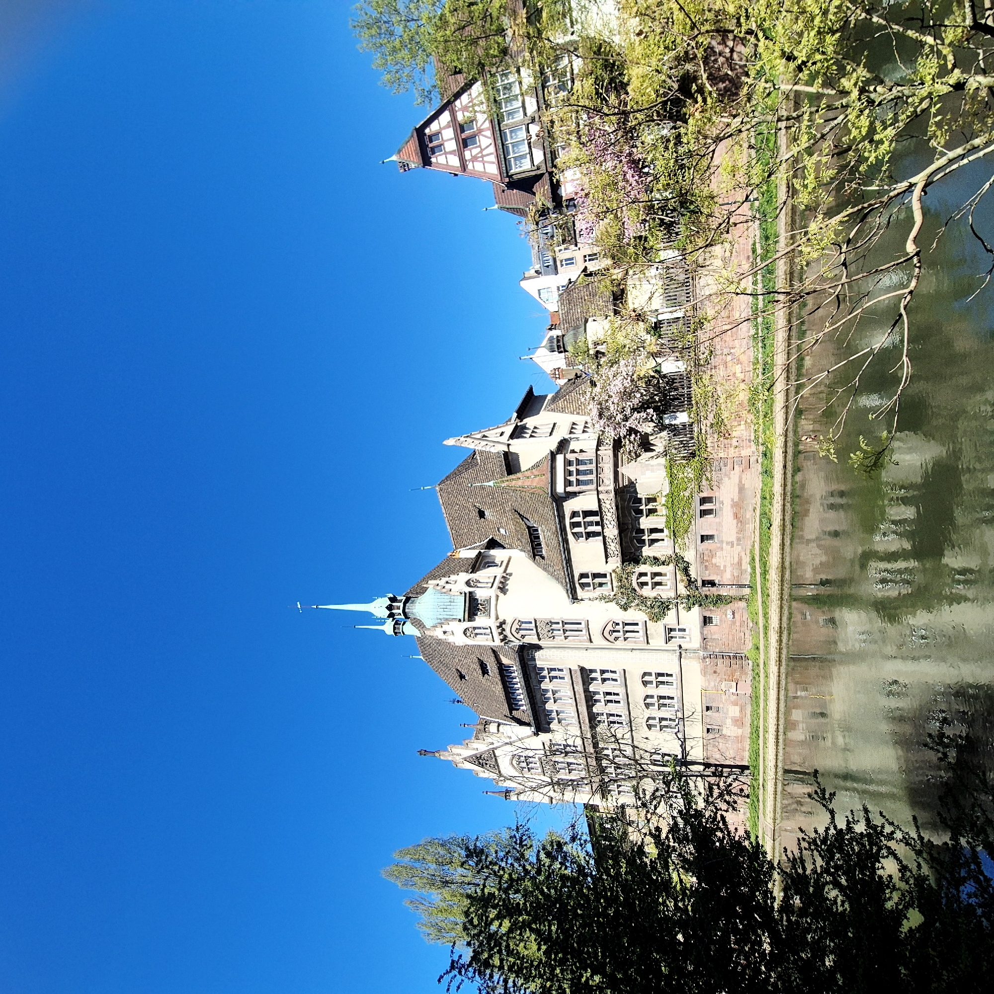 The image depicts a view in Strasbourg, France, with a river or canal running parallel to a row of buildings. The buildings showcase a variety of architectural styles, with some featuring half-timbered facades and others displaying more ornate details, including pointed roofs and turrets. Lush green trees and vegetation line the canal, and the reflections of the buildings are visible in the water. The sky is a clear blue. The location appears to be a historic district of the city.