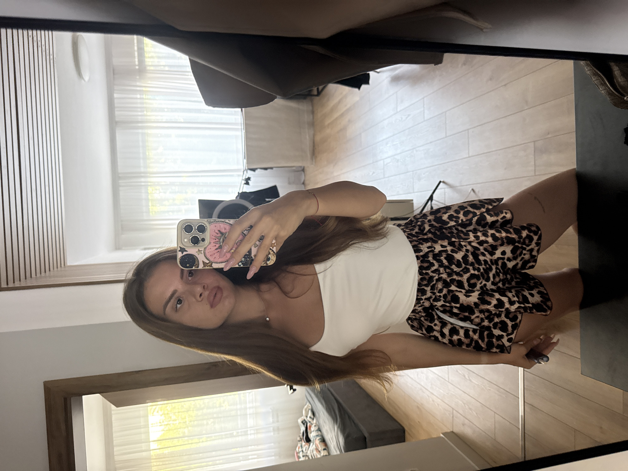 A young woman is taking a mirror selfie in a room. She is wearing a white tank top and leopard print shorts. She is holding a phone with a floral case in her right hand and a black object in her left hand. The room has wooden floors and white curtains. There is a bed with a grey duvet in the background. The woman has long brown hair and is wearing light makeup. The image is taken from a slightly low angle. There is no indication that this location is in Plovdiv, Bulgaria.