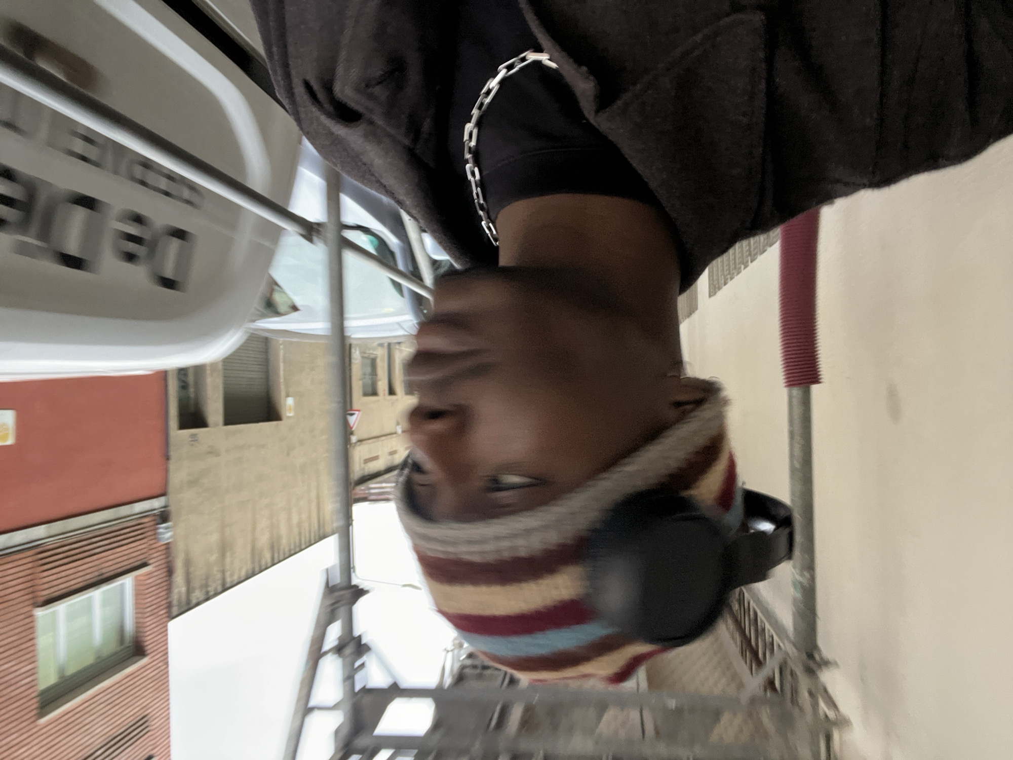 A person wearing headphones and a striped hat stands on scaffolding in Barcelona, Spain. The person is wearing a black coat with a silver chain. The background includes residential buildings and a vehicle with writing on its side.
