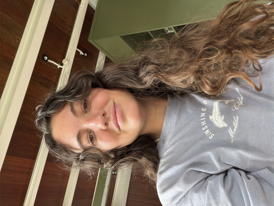 A young woman with curly brown hair, styled with gray highlights, smiles gently at the camera. She's wearing a light blue long-sleeved shirt adorned with the words "Sunshine Beach, Malibu, California" encircling a drawing of a dolphin. The background shows a wooden ceiling, white beams, and a light green cabinet partially visible. The image is taken at a slightly tilted angle, creating an interesting perspective.