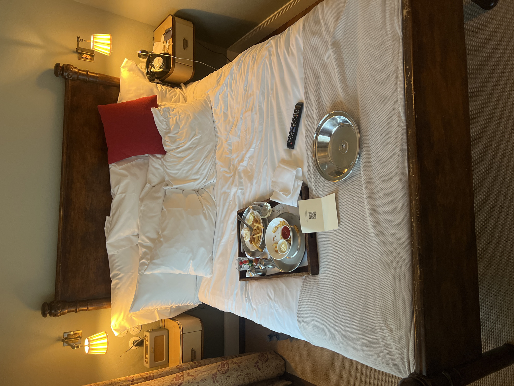The image depicts a hotel room interior. The focus is on a bed with a dark wooden frame and white linens. A red pillow is placed near the headboard. On the bed, there is a remote control, a silver tray, and a food tray with dishes and a white paper with a QR code. The room has a warm lighting, with bedside lamps illuminating the area. The walls are painted a light green color. There is a piece of luggage next to one side of the bed. A carpeted floor is visible. The location cannot be determined by the image.