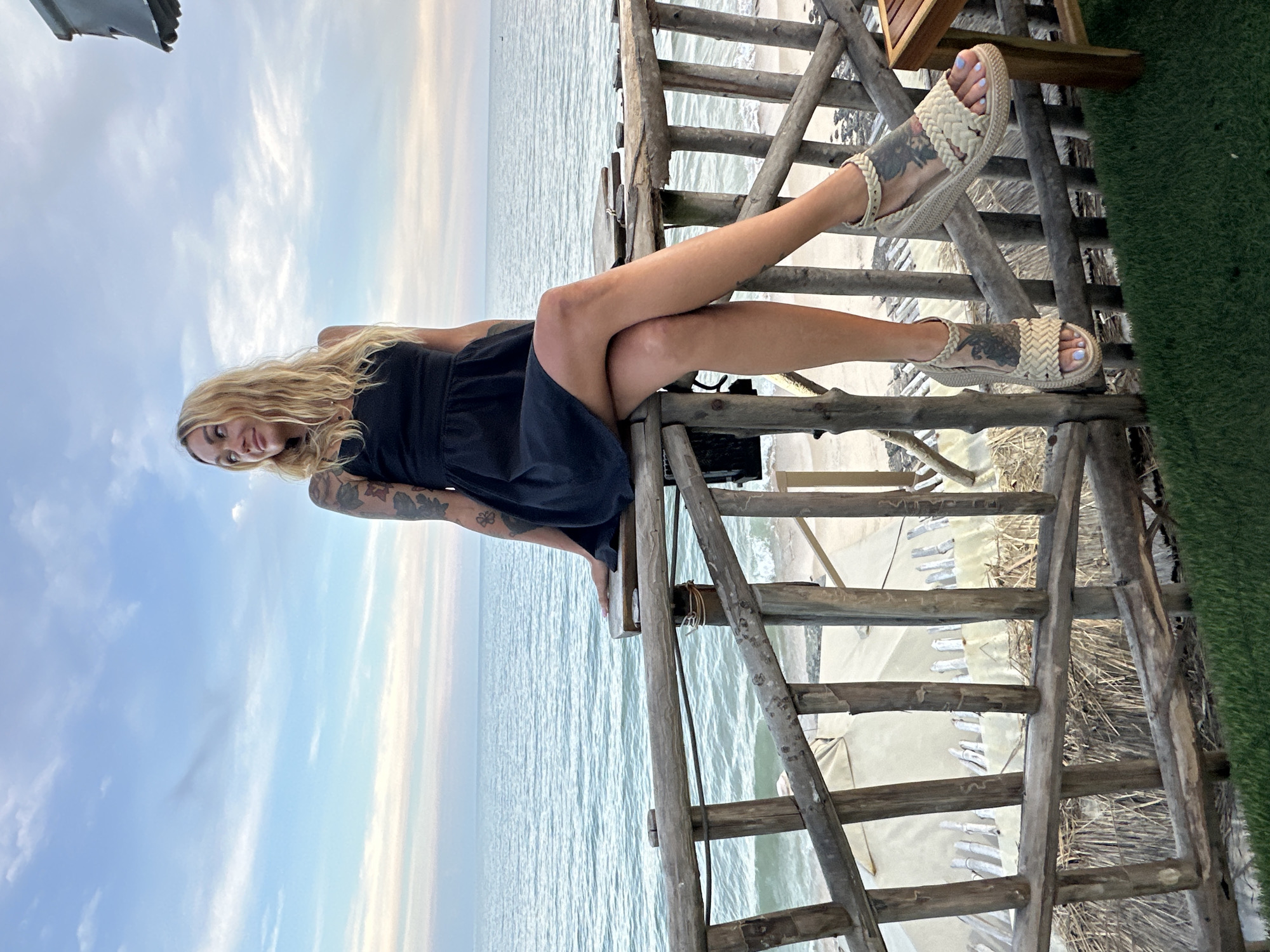 The image shows a woman sitting on a wooden railing overlooking the sea. She has blonde hair, is wearing a black dress and sandals, and has tattoos on her arms and legs. The setting appears to be a beachside location, likely in Phu Thuy Ward, Vietnam, given the typical structures and vegetation found in such areas. The sea is calm with gentle waves, and the sky is partly cloudy with soft colors indicating either sunrise or sunset. In the background, there are some light-colored structures with thatched roofs, suggesting a resort or beach bar. The overall atmosphere is relaxed and scenic.